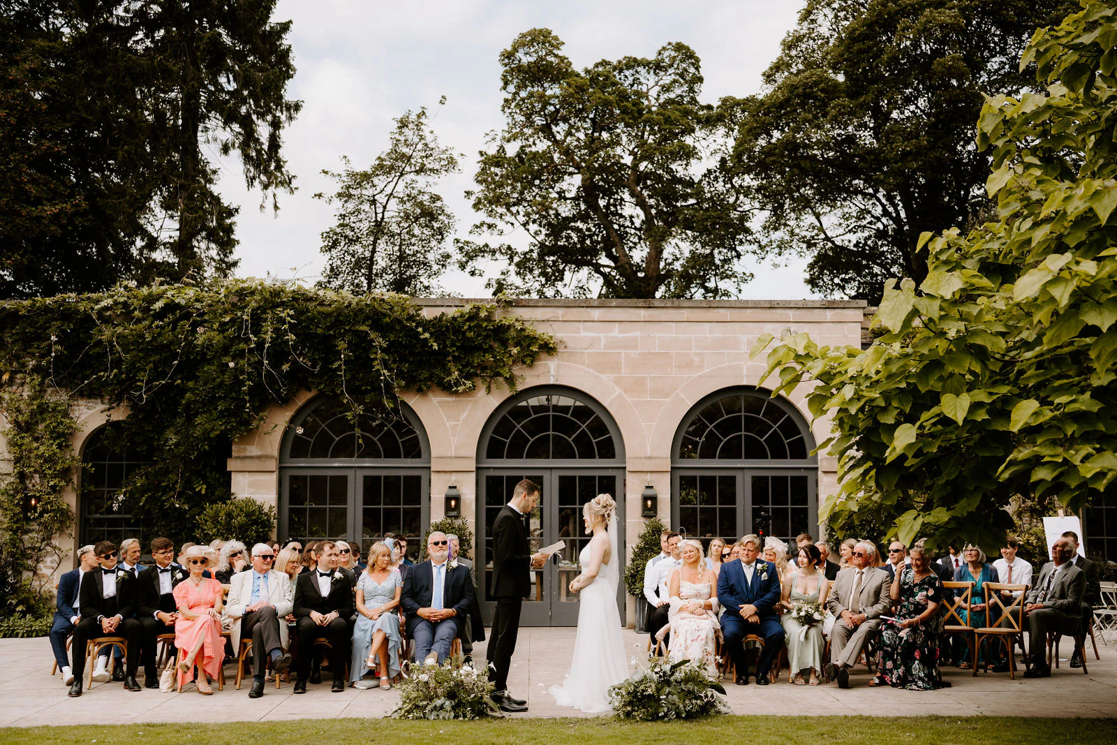 Wedding guests seated outdoors in front of an arched stone building while a couple stand at the front for their ceremony under tall trees