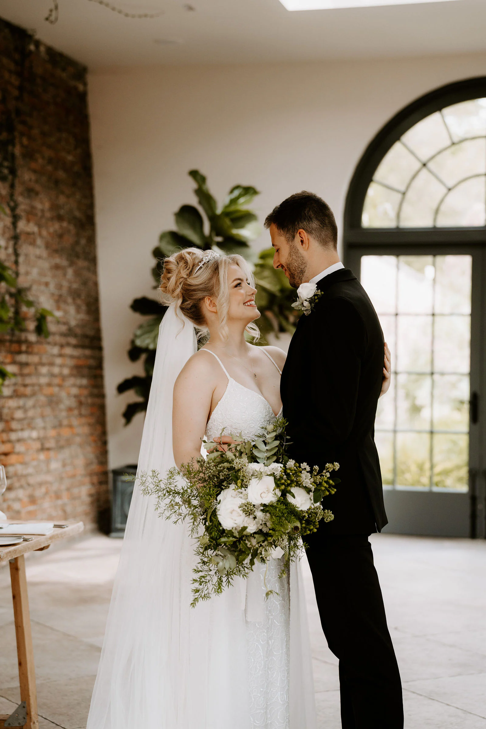 A bride holding a bouquet of white flowers and a groom stand together in a bright room with tall arched windows