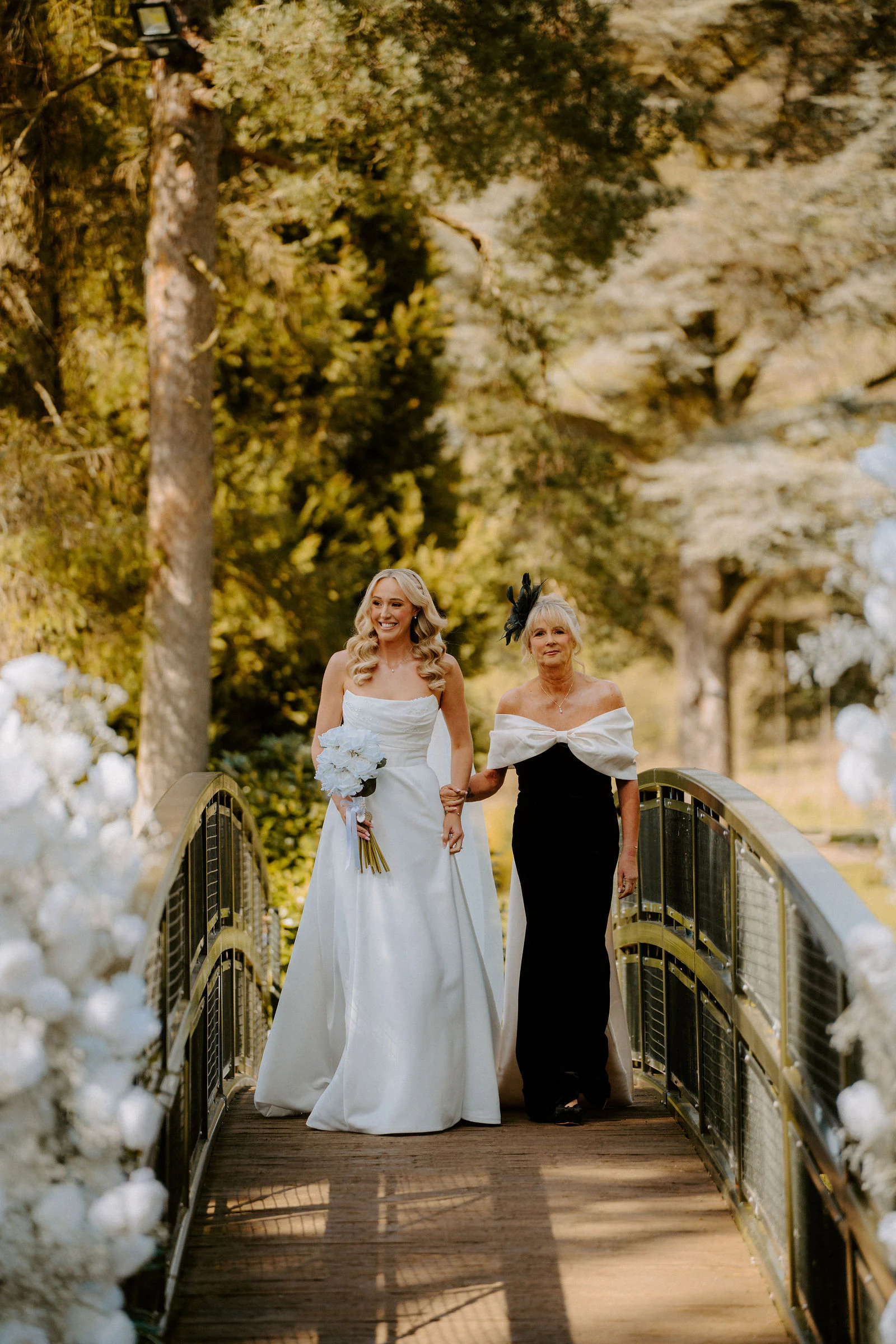 A bride and an older woman in black walk across a small bridge lined with white flowers in the trees