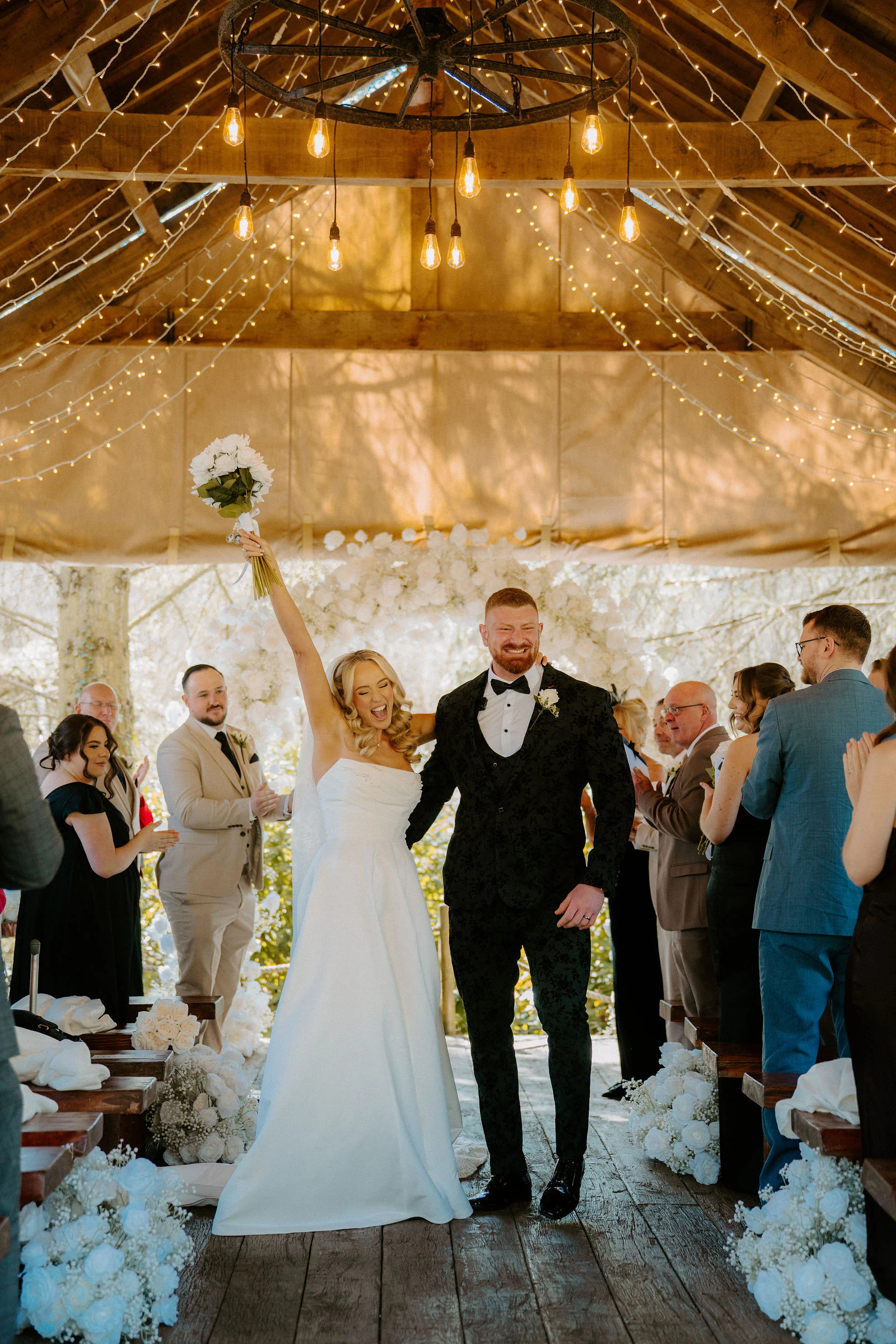 A bride raises her bouquet as she and her groom walk down the aisle beneath a timber canopy hung with fairy lights