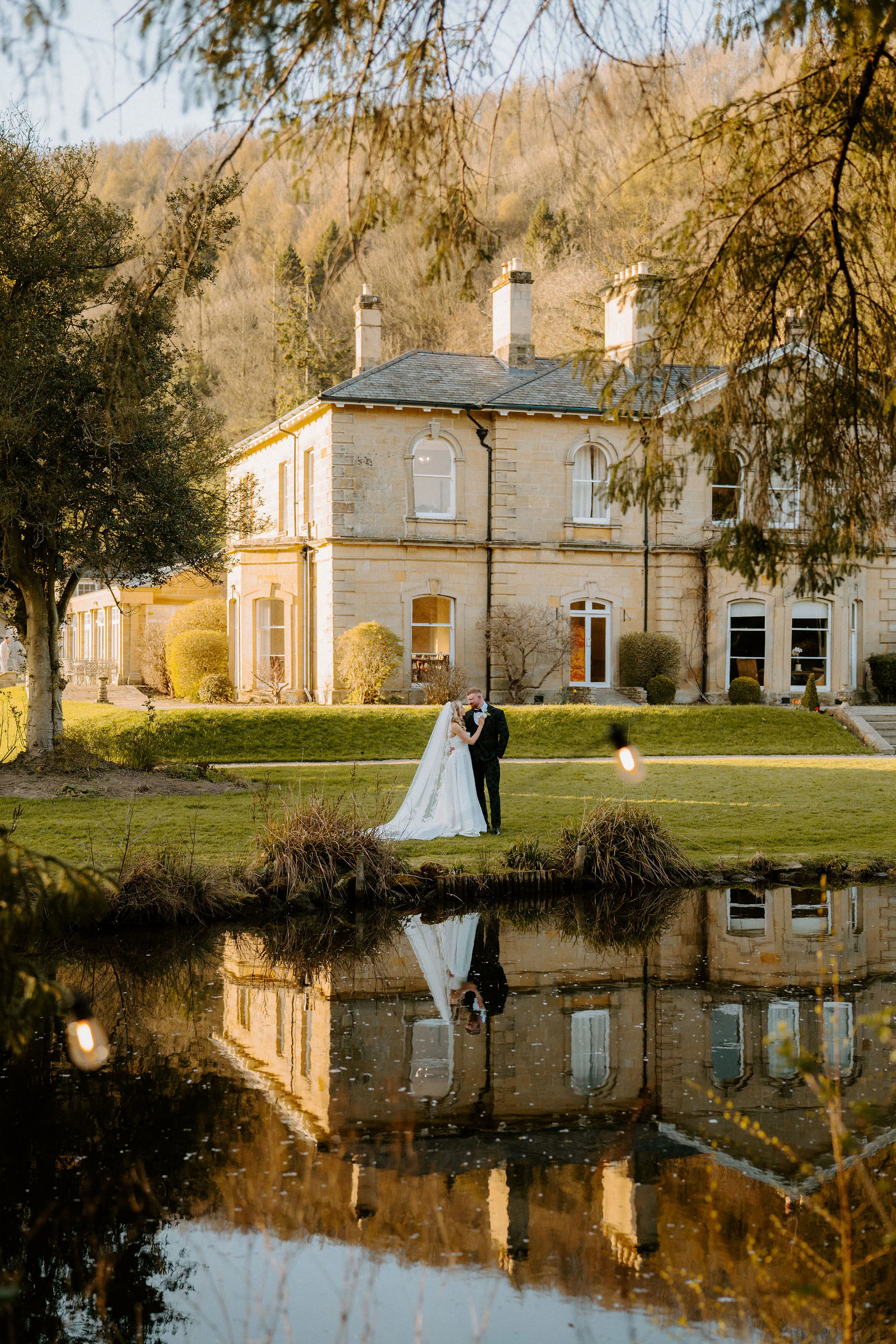 A bride and groom embrace on a lawn in front of a stone house with its reflection in a pond