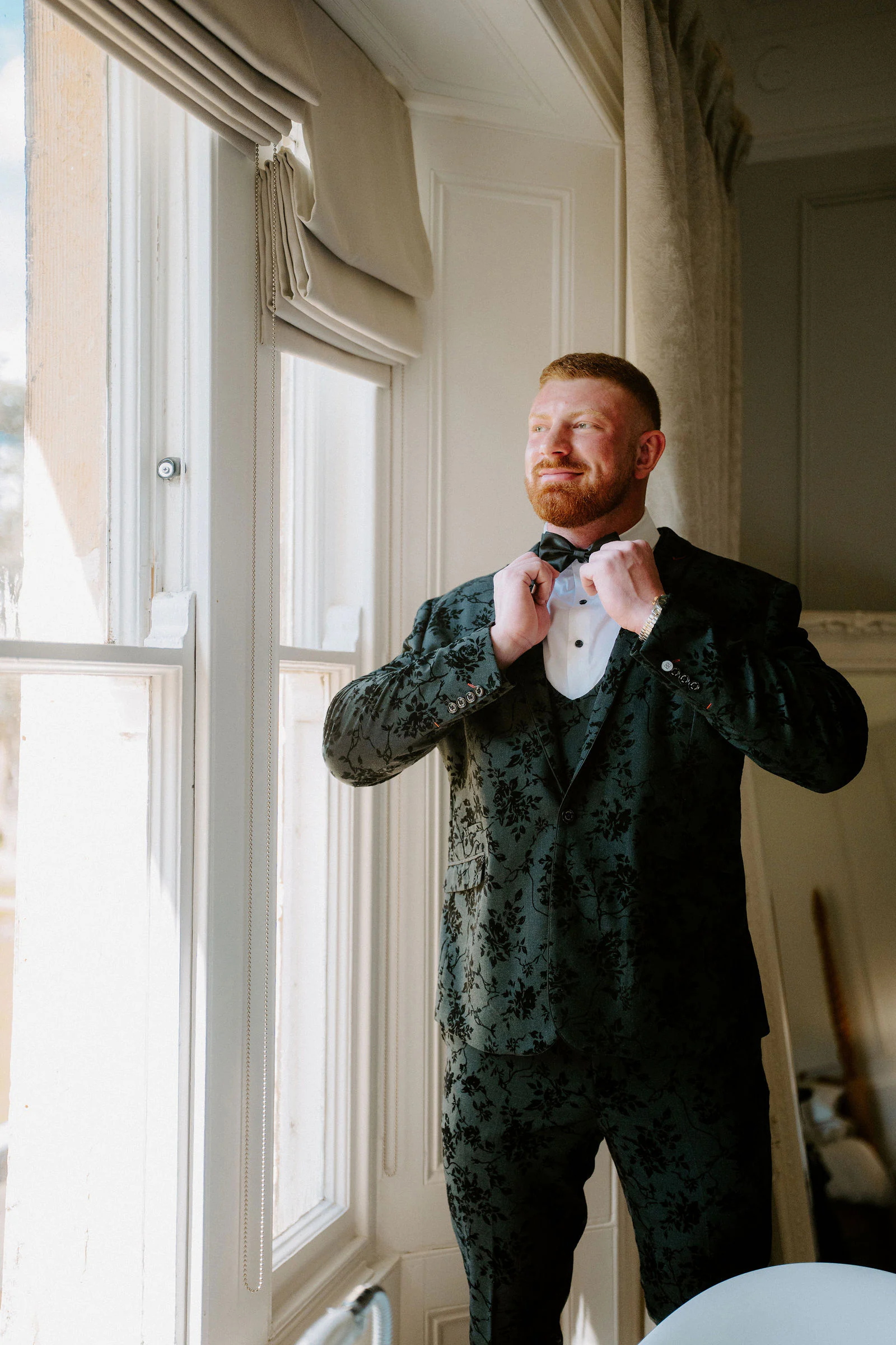 A groom in a dark green patterned jacket adjusts his bow tie beside a tall sash window