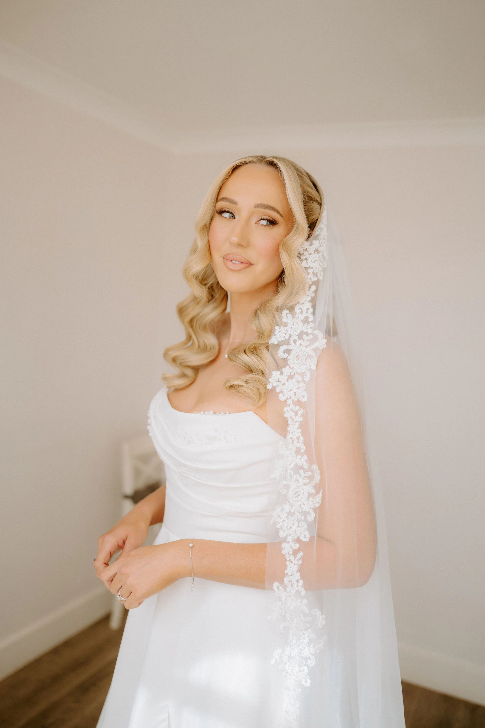A bride in a lace-edged veil glances over her shoulder in a plain bright room