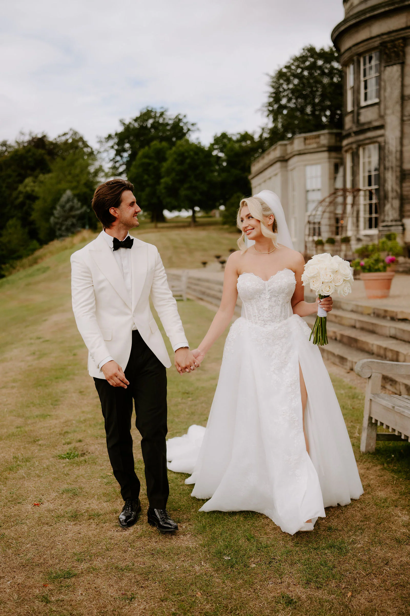 A groom in a white dinner jacket and black trousers and a bride in a strapless gown with a thigh-high split walk hand in hand across dry grass, with a stone wing of the hall and a wooden bench behind them