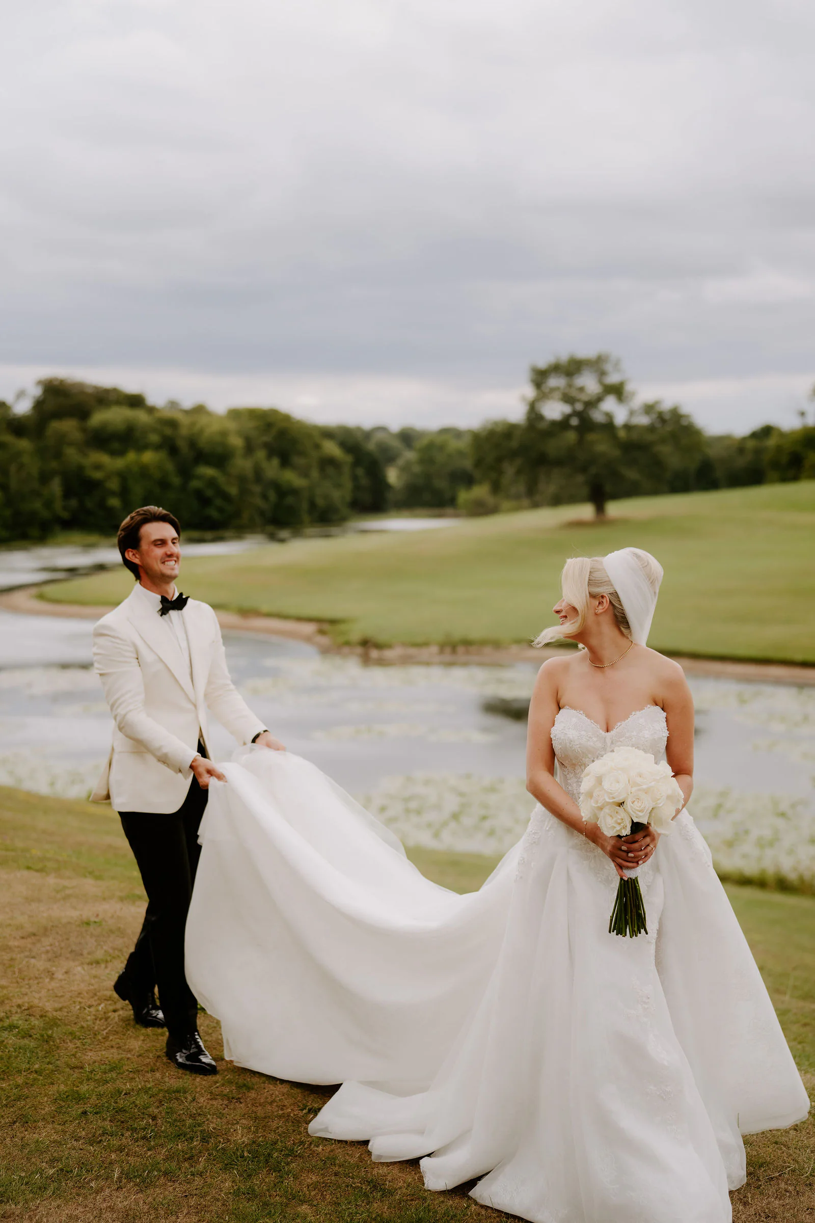 A groom in a white dinner jacket holds up the long train of his bride's gown as she looks back over her shoulder, with a lake with lily pads and a wide green lawn behind them