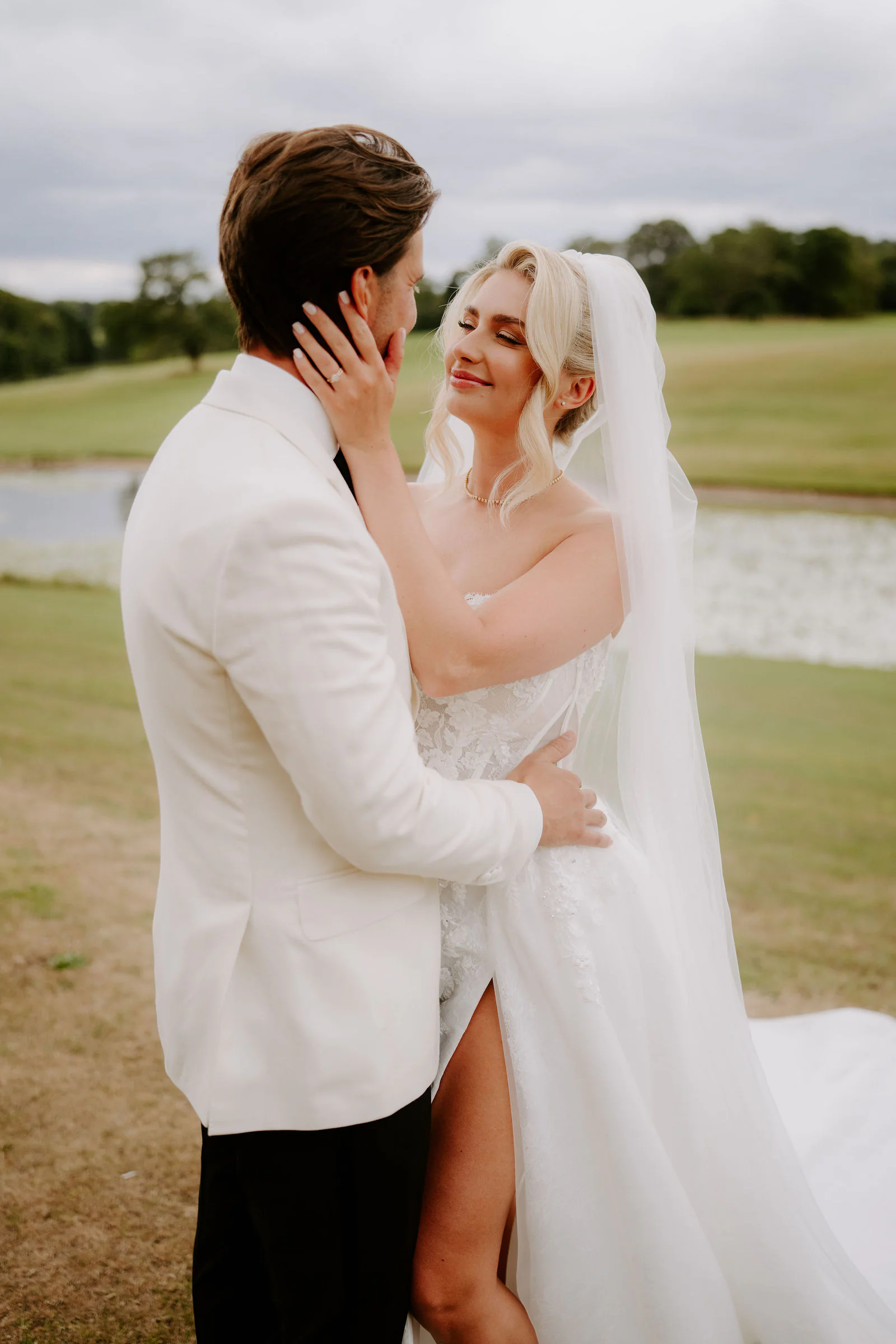 A groom in a white dinner jacket holds a bride's face as they embrace, her long veil blowing, with a lake and green parkland behind them under a grey sky