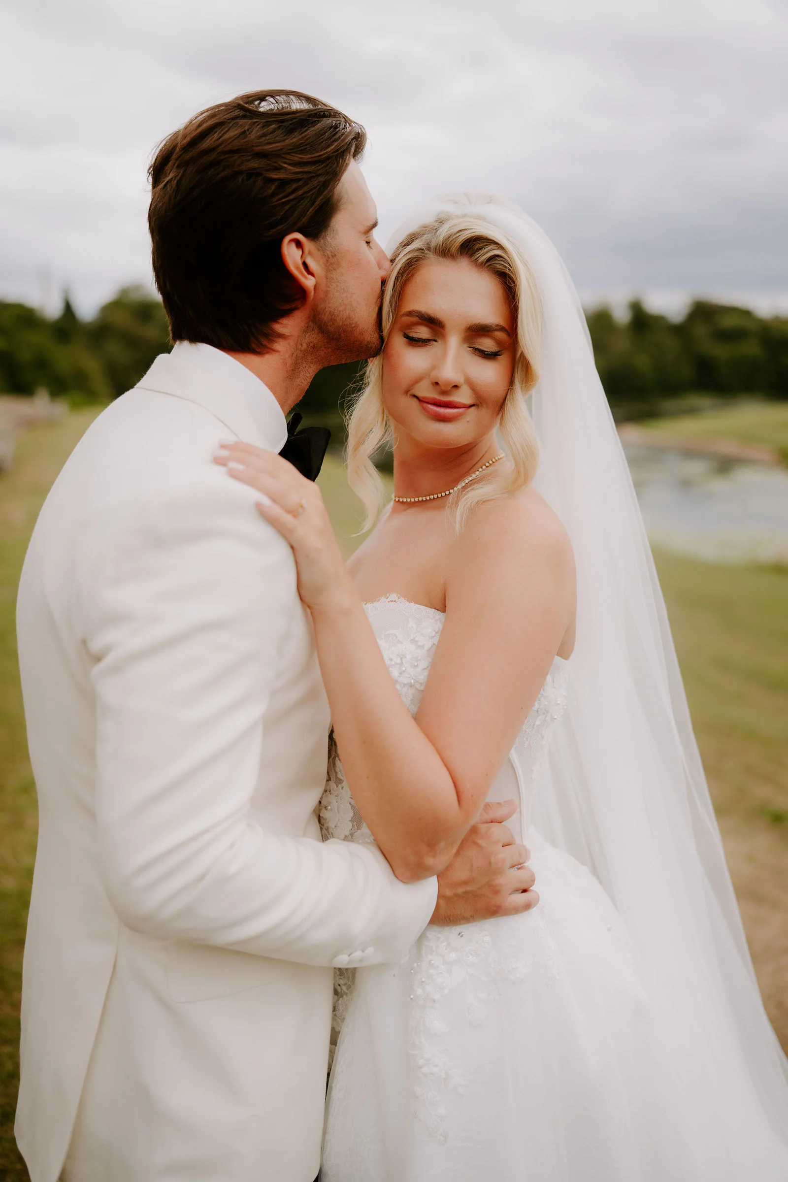 A close portrait of a groom in a white dinner jacket kissing his bride on the forehead as she closes her eyes, with her veil and a lake behind them