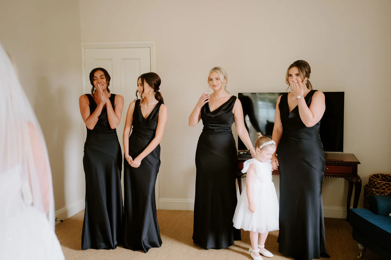 Four bridesmaids in black satin dresses and a small flower girl in white react with their hands to their faces, as the edge of a bride's veil shows at the left of the frame