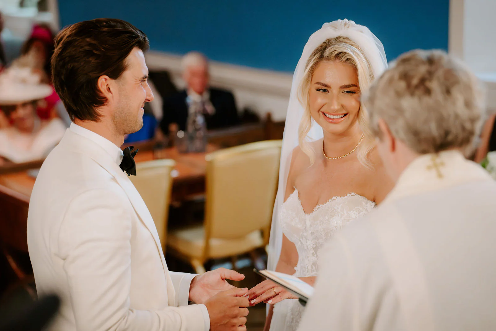 A bride in a veil smiles at her groom in a white dinner jacket as he holds her hand during the ring exchange, with a person in cream vestments in the foreground and seated guests behind
