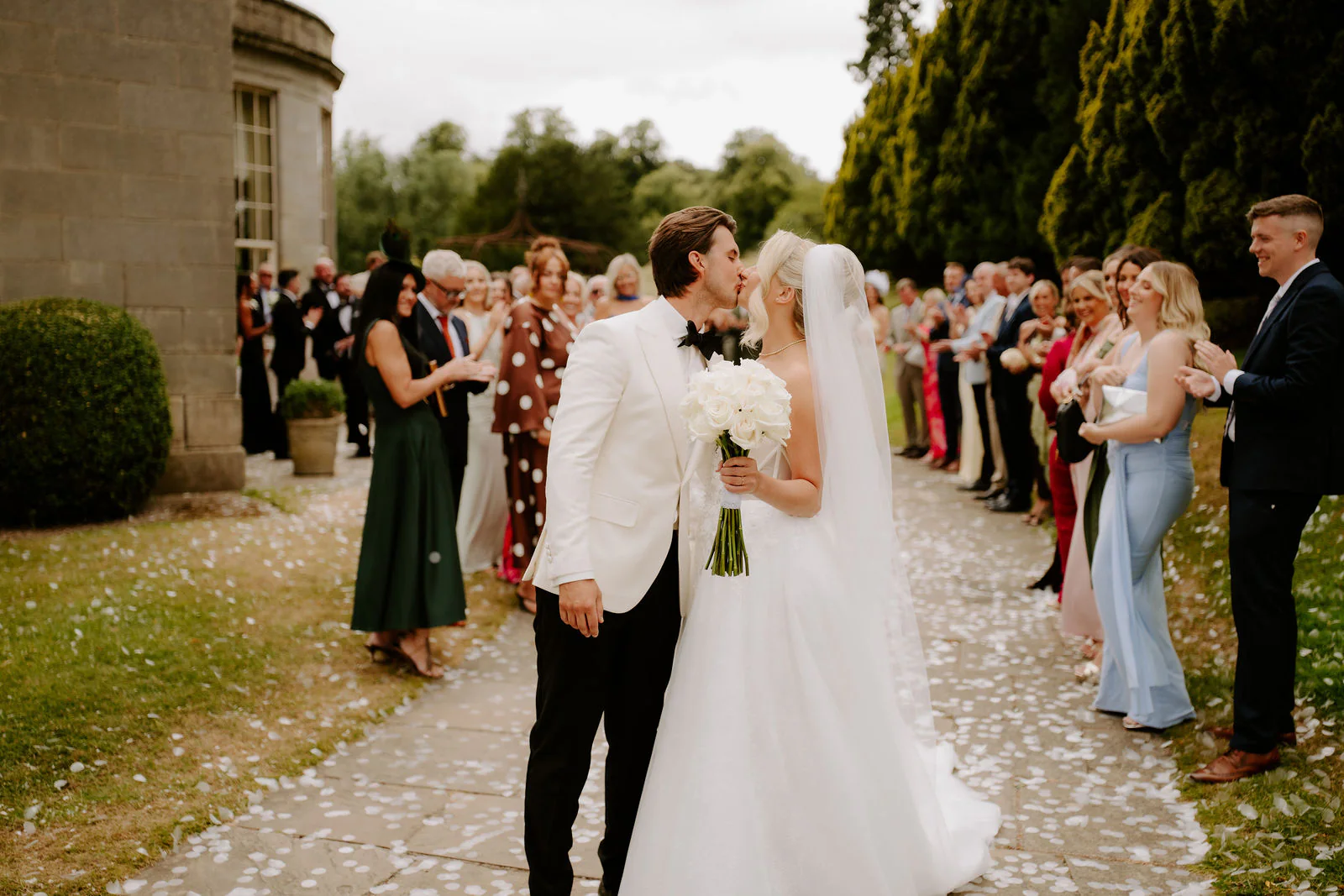 A groom in a white dinner jacket kisses a bride with a long veil on a stone path scattered with white petals, with guests lined up on both sides and a curved stone wing of the hall at the left