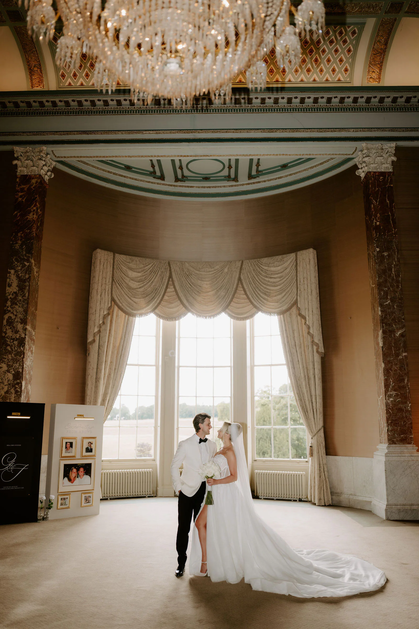 A bride in a long train and a groom in a white dinner jacket look at each other in a grand room with a crystal chandelier, marble columns, a curtained bay window and a board of framed family photographs