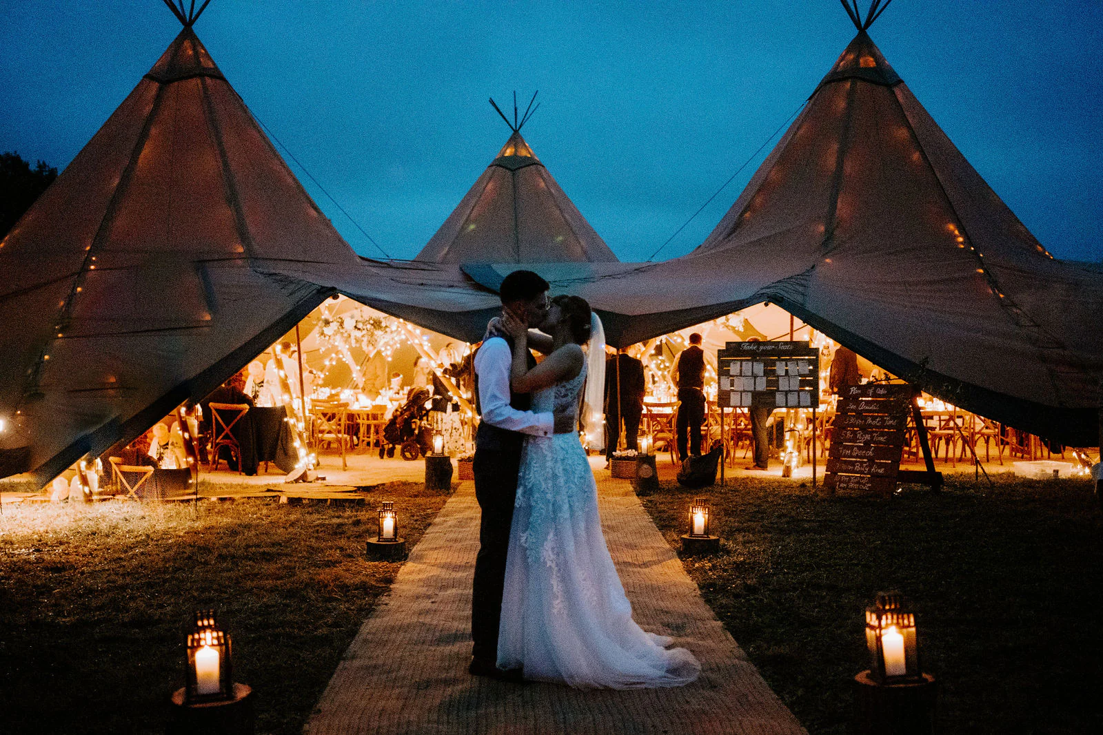 A bride and groom kiss on a lantern-lit path in front of large illuminated tipi tents at dusk