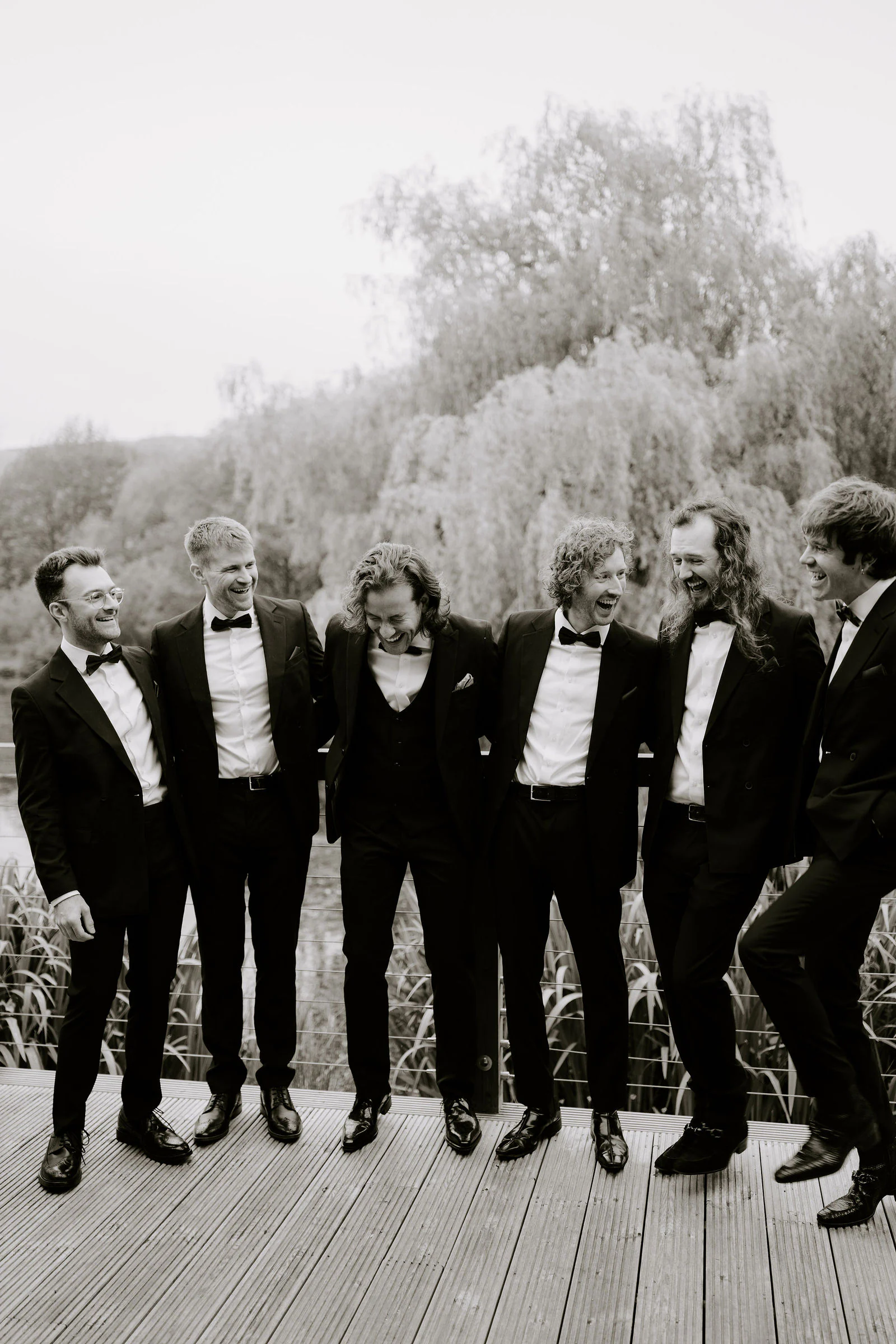 A bridal party of groomsmen in black tuxedos laughing together on a wooden deck beside willow trees and water, in black and white