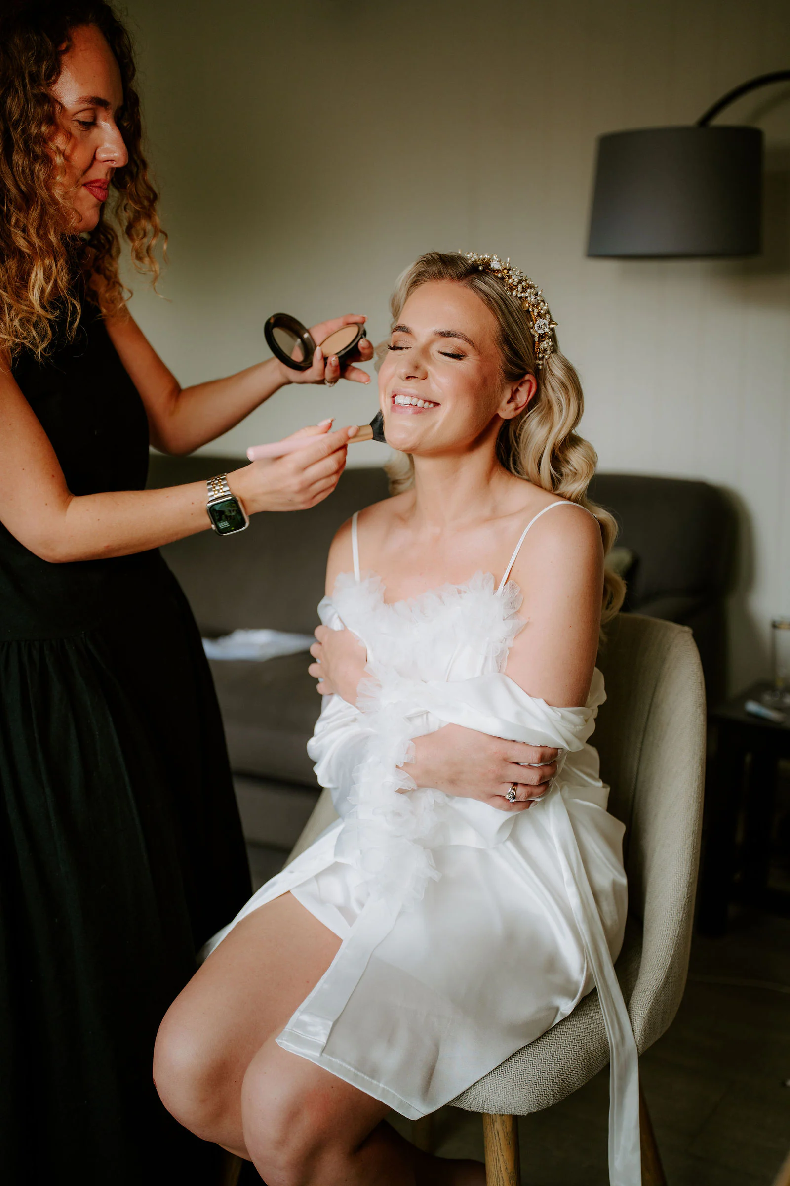 A make-up artist brushes the cheek of a bride in a white feathered robe in a softly lit room