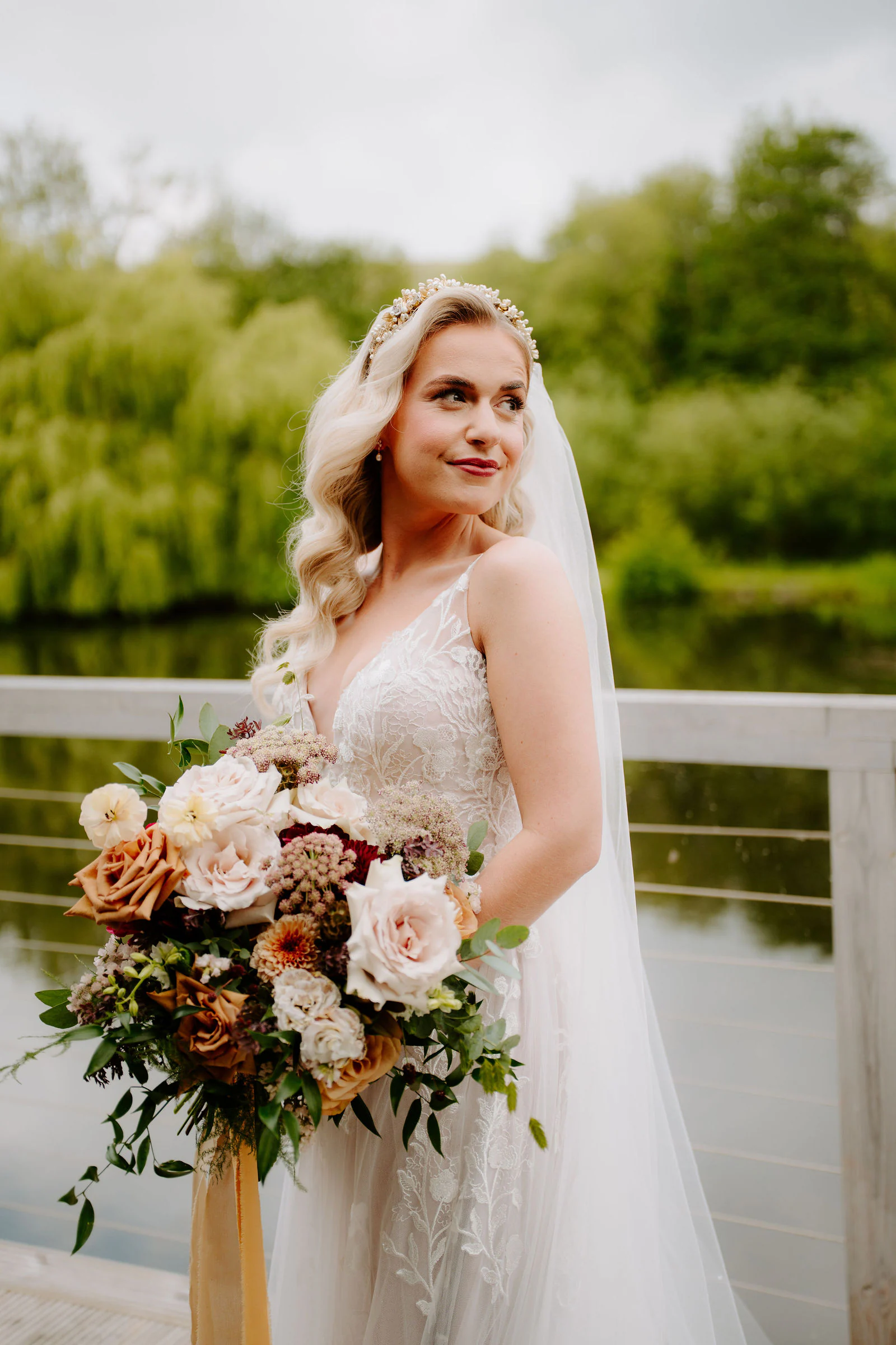 A bride in a floral crown holds a warm-toned bouquet on a balcony beside a lake and willows