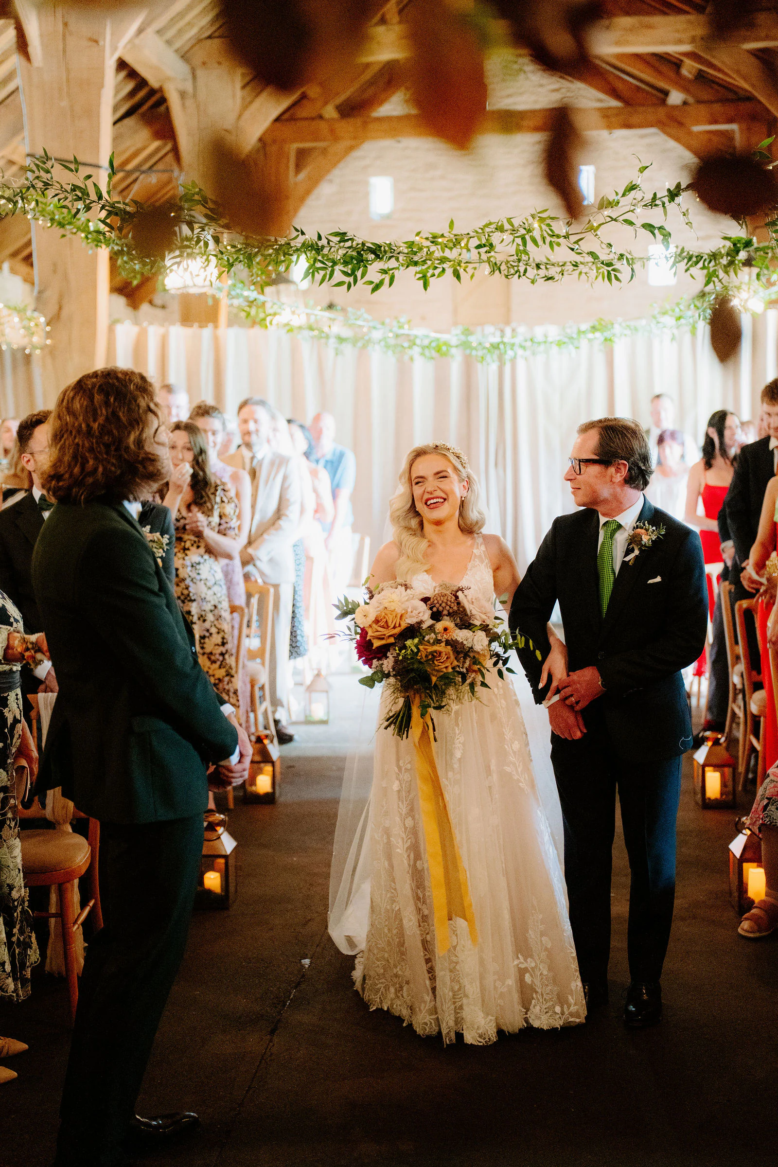 A bride walks down the aisle beside an older man under timber beams hung with greenery as guests smile