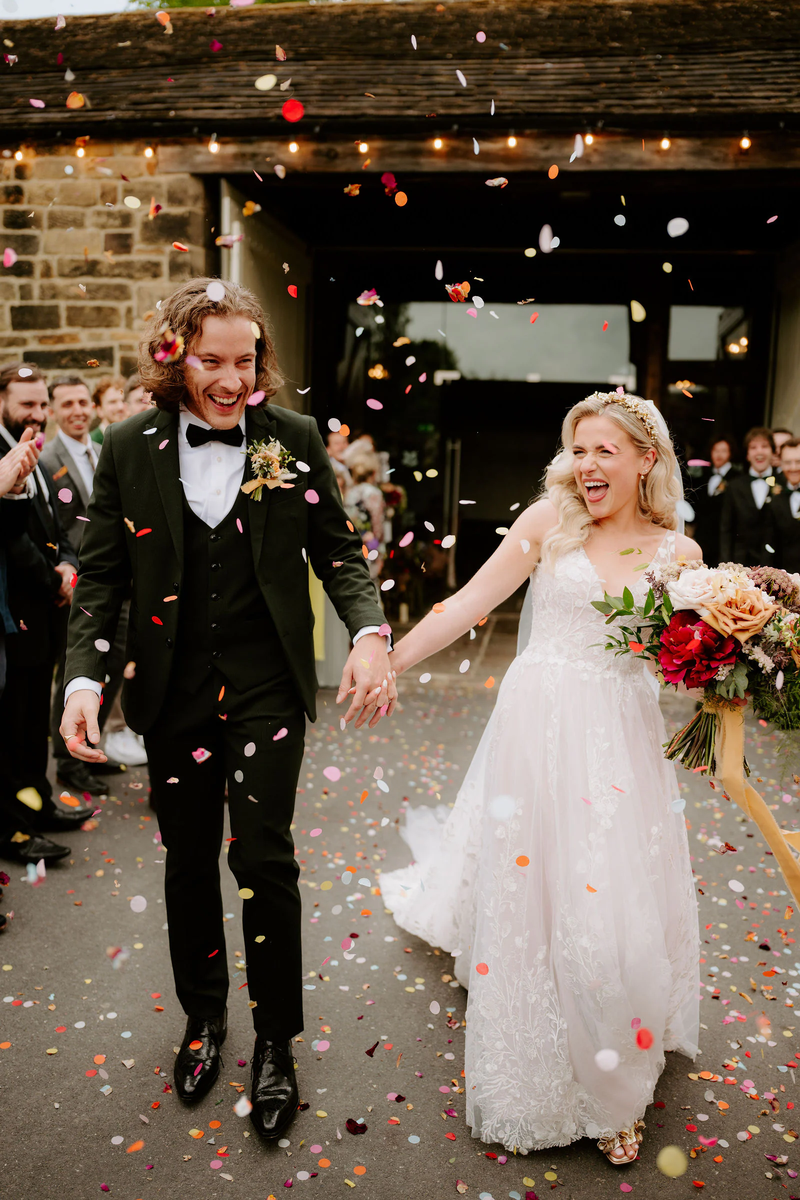 A couple walk laughing through a shower of confetti out of a stone barn lit by fairy lights