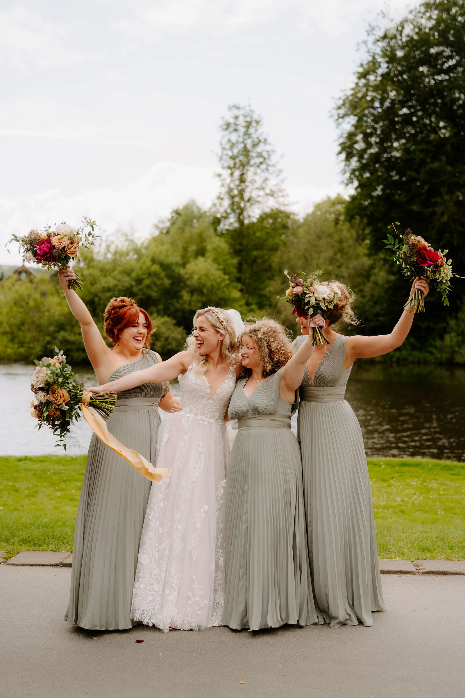 Bridesmaids in sage green dresses and the bride throw up their bouquets beside a lake
