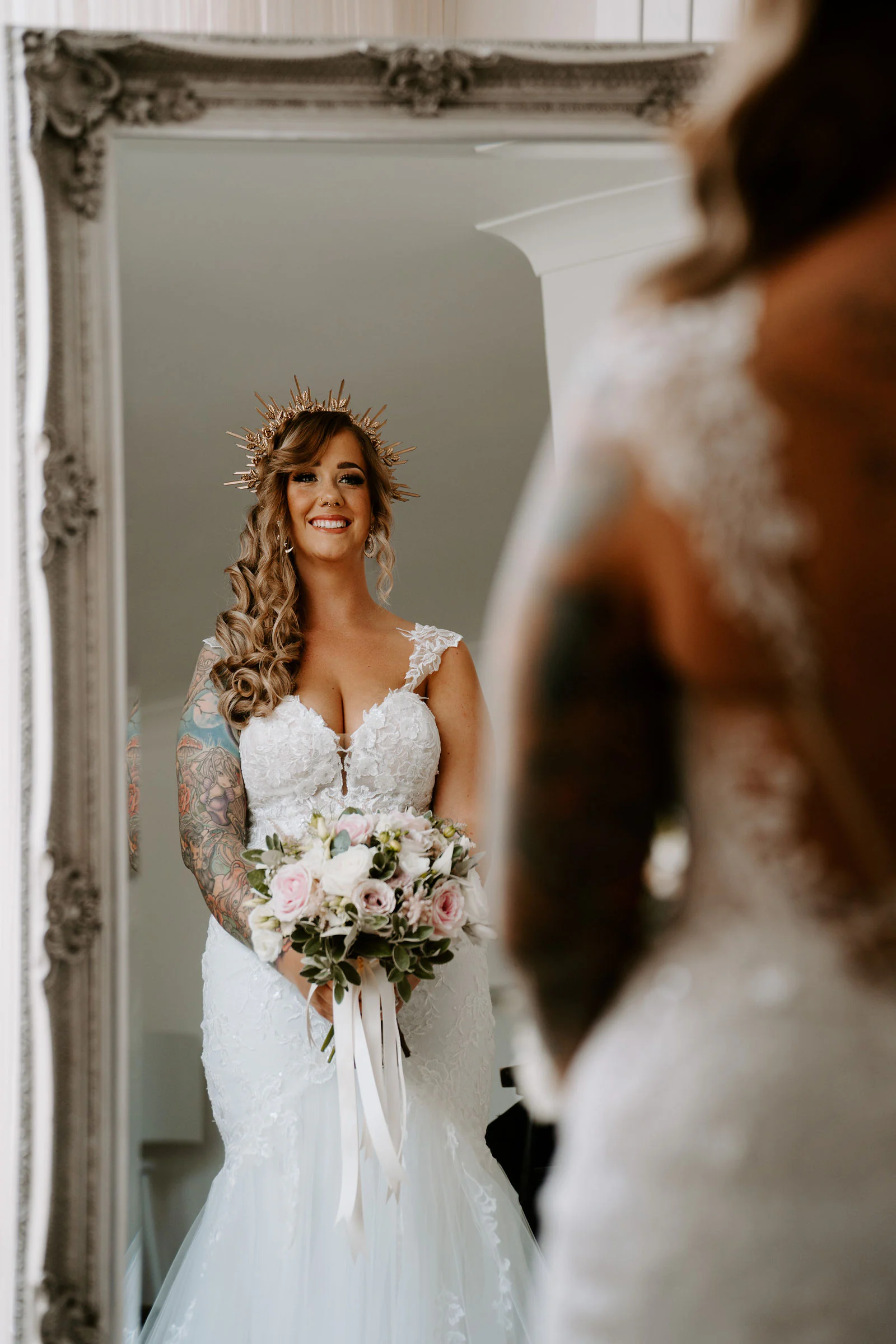 A bride in a gold sunburst crown holds a bouquet of pink and white roses in front of a large ornate mirror