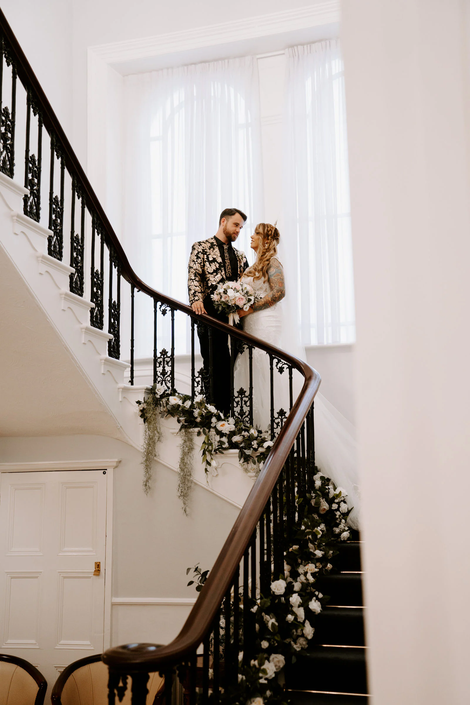 A bride and groom stand on a white staircase with a black iron balustrade wound with greenery and white flowers