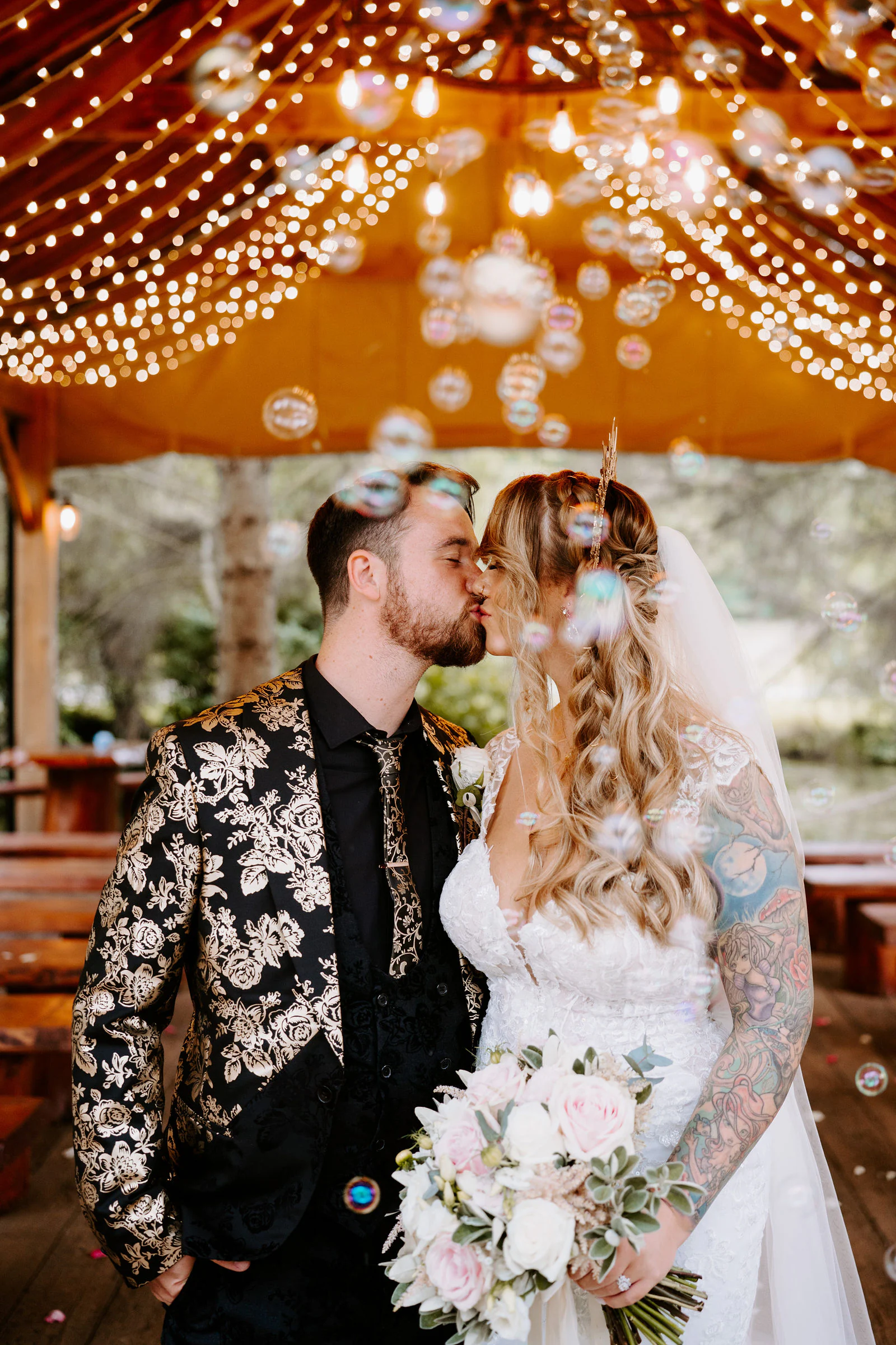 A bride and groom kiss as soap bubbles float around them beneath a canopy of fairy lights