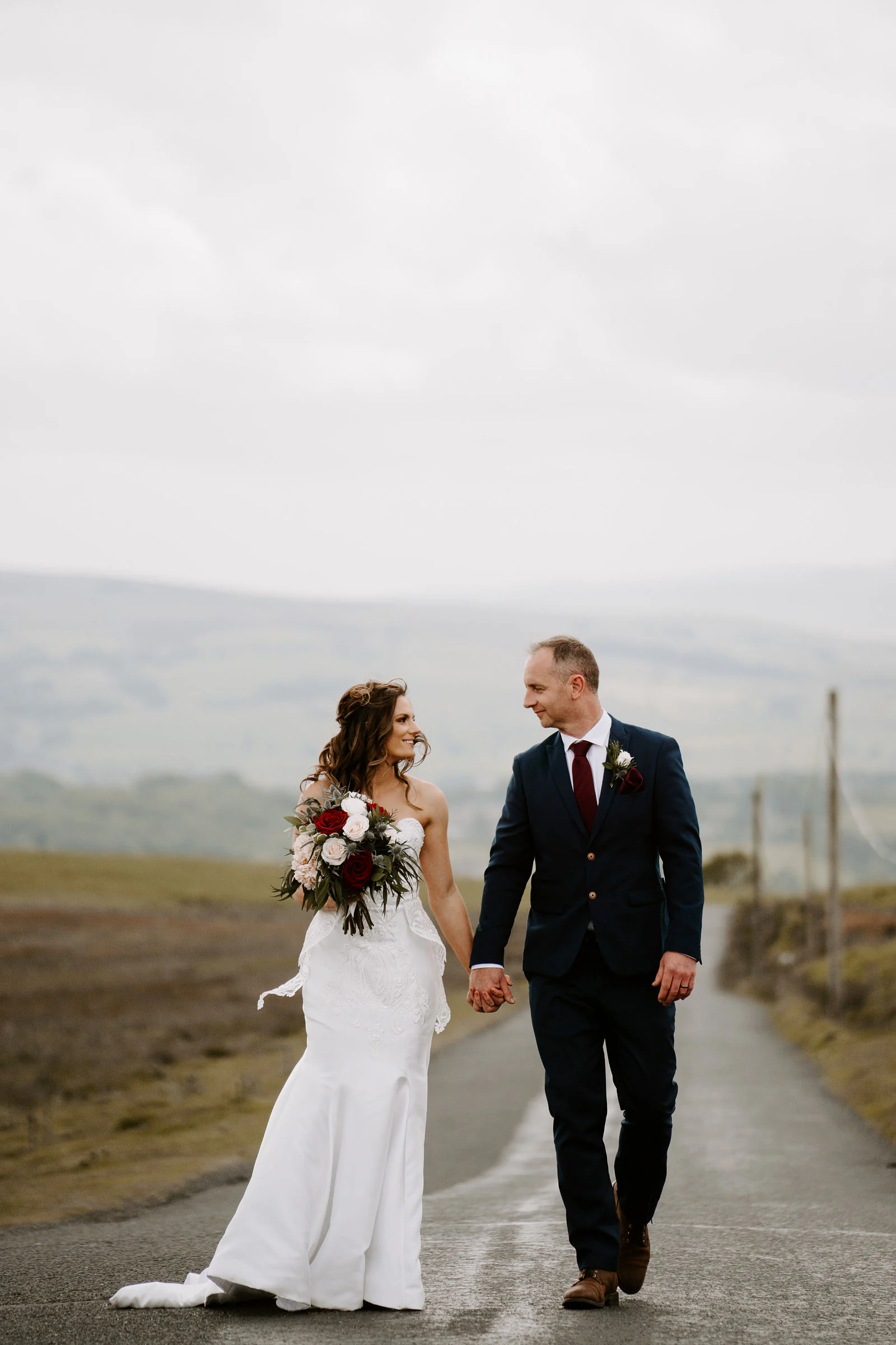 A bride and groom hold hands and walk along a quiet road across open moorland under a cloudy sky