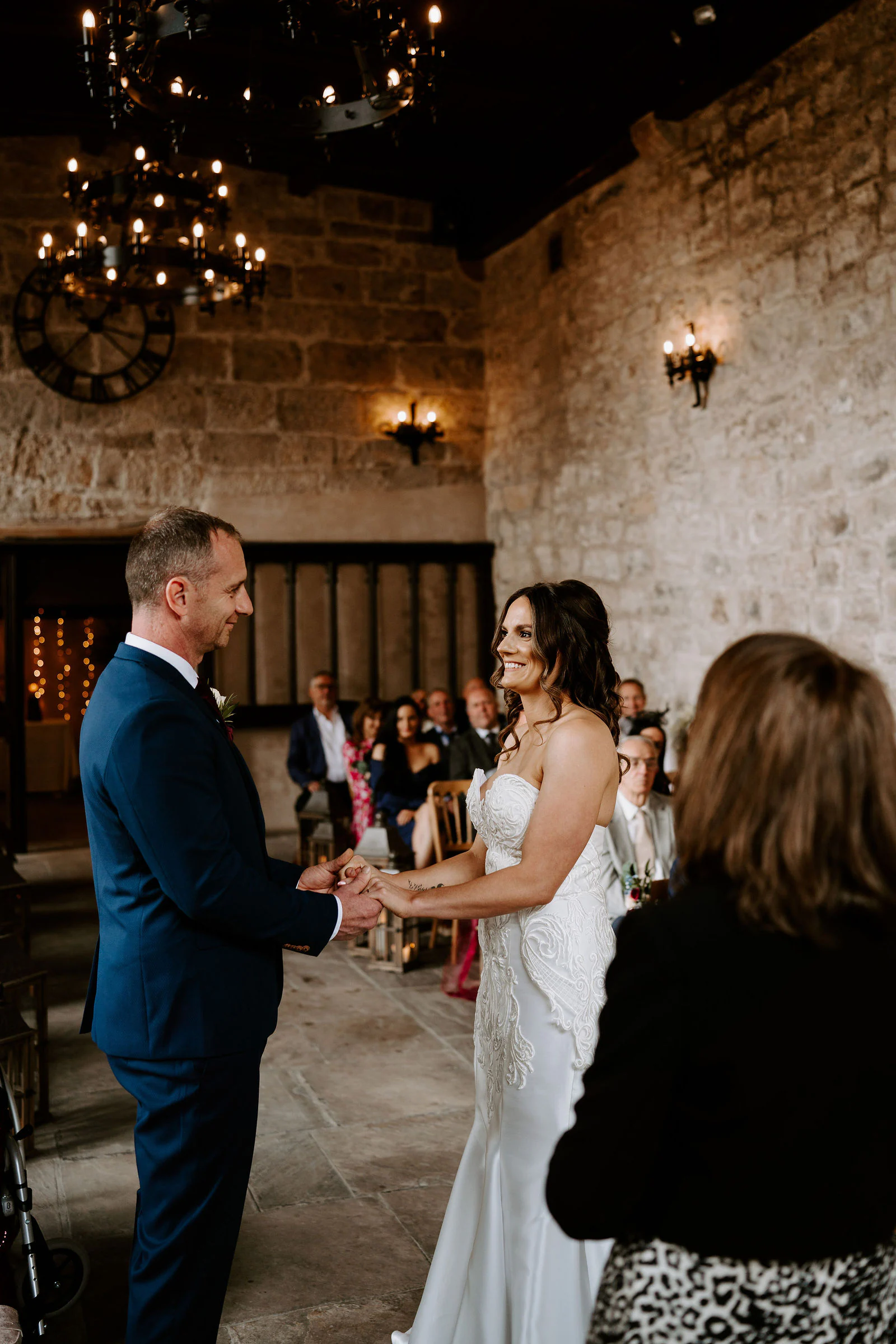 A bride and groom hold hands during their ceremony in a stone hall lit by iron chandeliers
