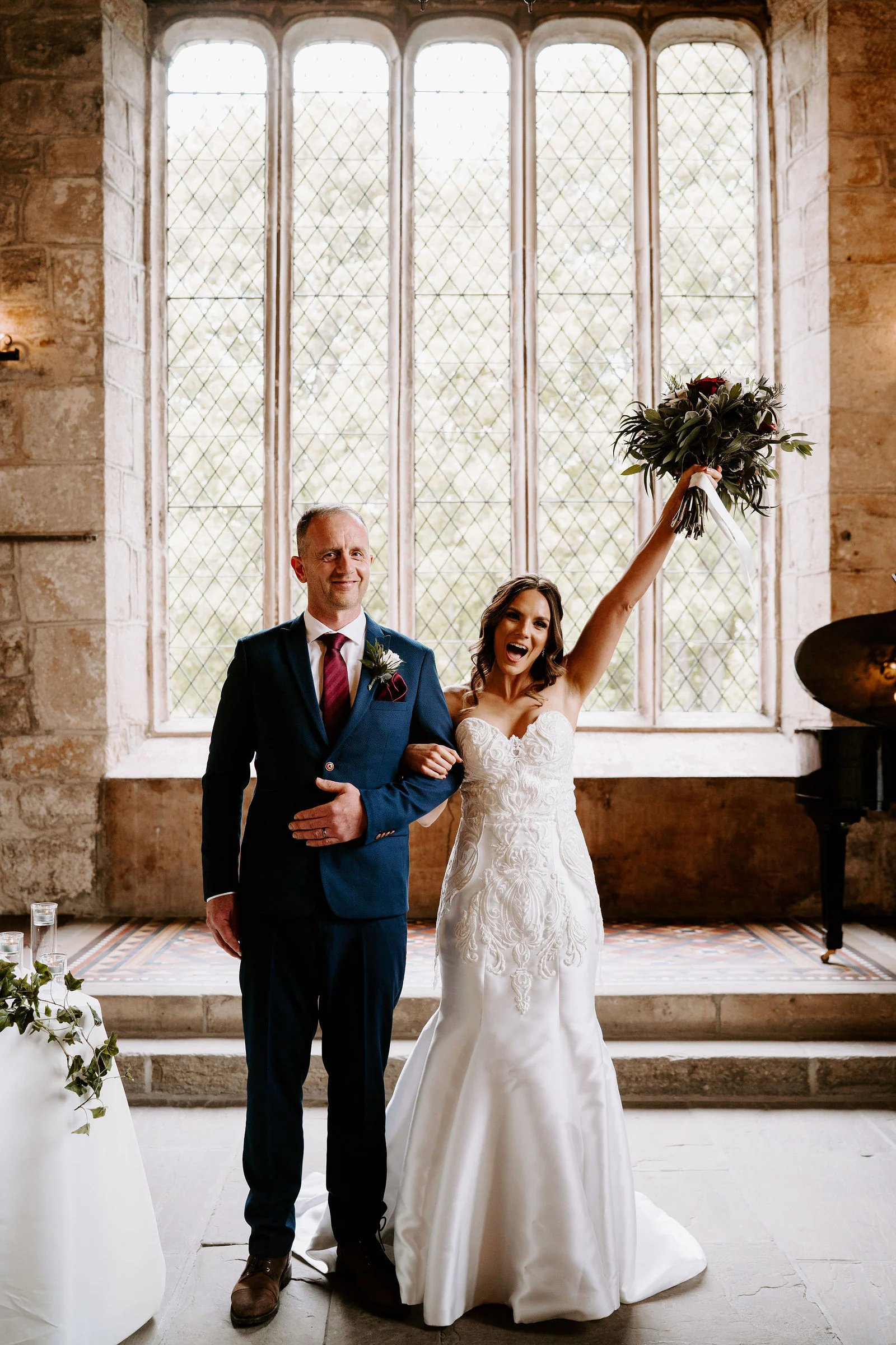 A bride raises her bouquet and cheers beside her groom in front of a tall stone window with leaded panes