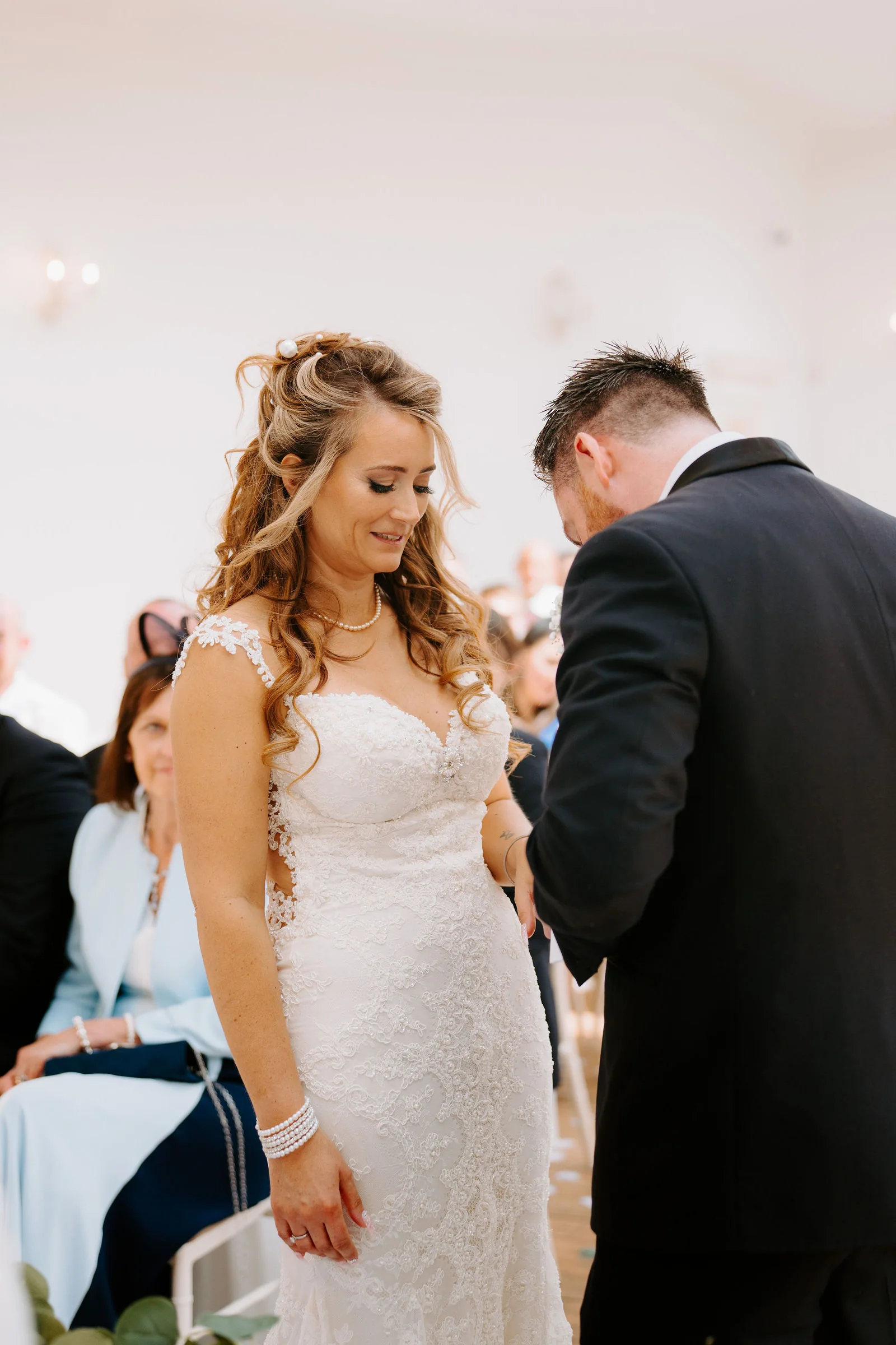A smiling bride looks down as her groom bows his head beside her in a pale ceremony room, with guests seated around them