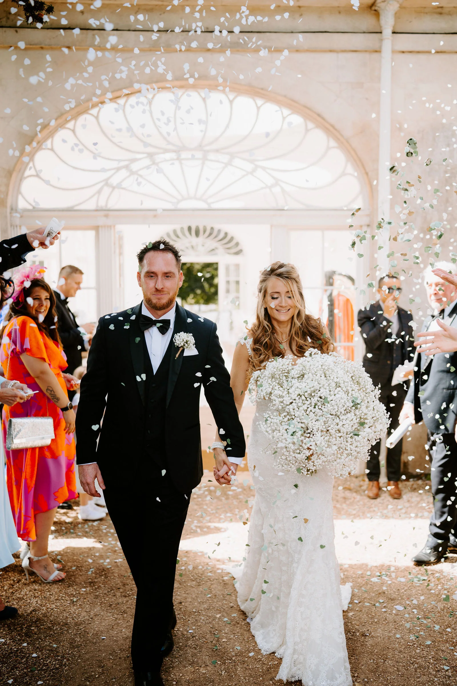A bride carrying a large bouquet of gypsophila and a groom in black tie walk out under an arched conservatory window as confetti falls, with guests either side