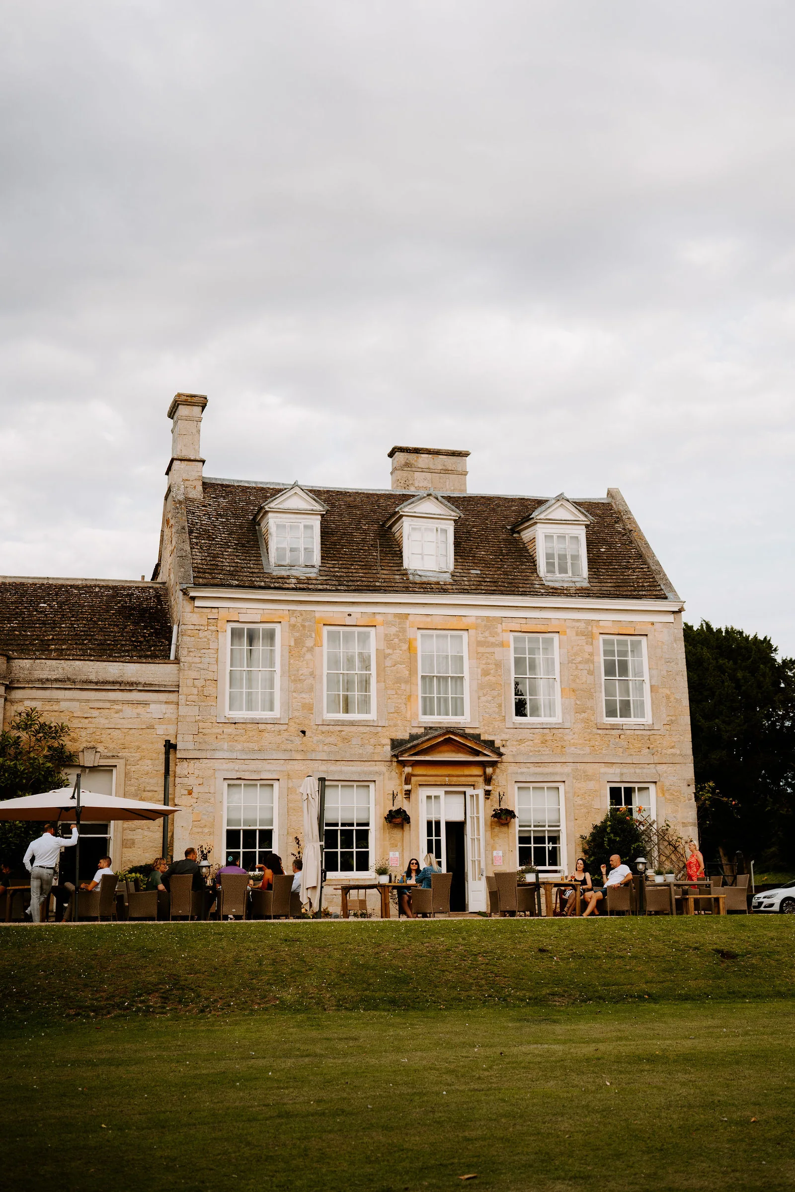 A stone manor house with dormer windows beyond a lawn, with guests seated outside on the terrace