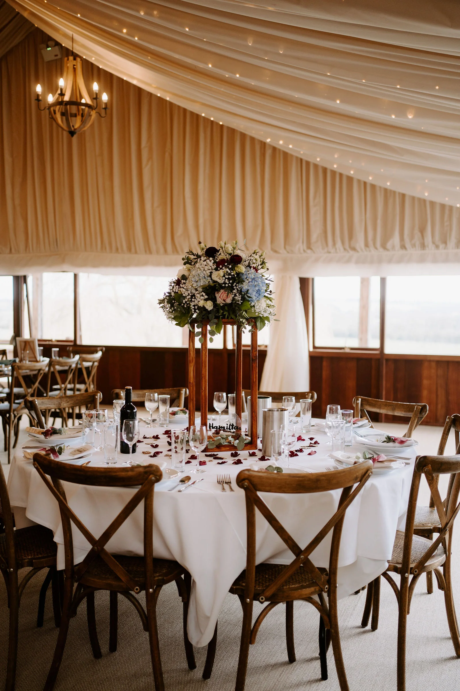 A round table laid for dinner with cross-back wooden chairs and a tall floral centrepiece inside a marquee lined with draped cream fabric and fairy lights, with a chandelier above