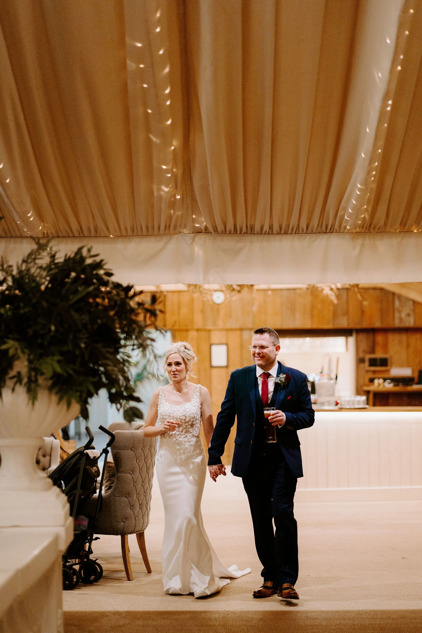 A bride in a fitted gown and a groom in a navy suit with a red tie, holding a drink, walk hand in hand through a marquee with a draped cream ceiling, a bar and wood panelling behind them