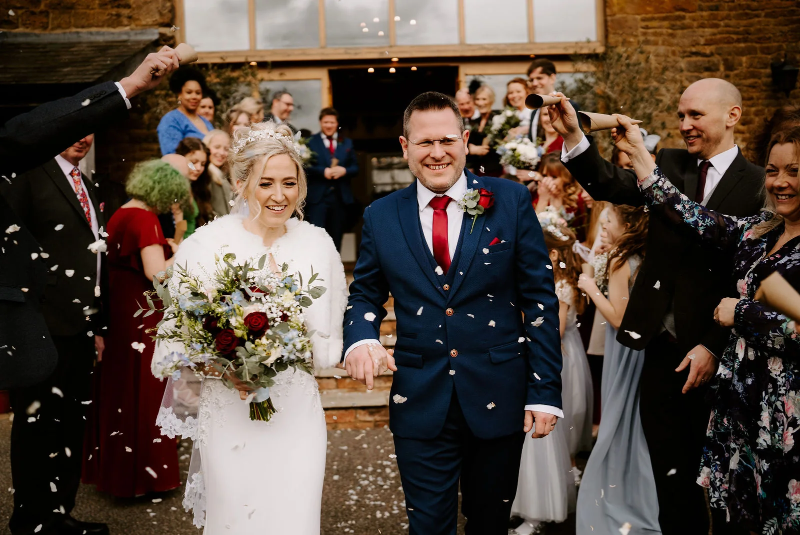 A bride in a white fur-trimmed wrap holding a bouquet and a groom in a navy suit with a red tie walk through guests throwing confetti outside a brick building