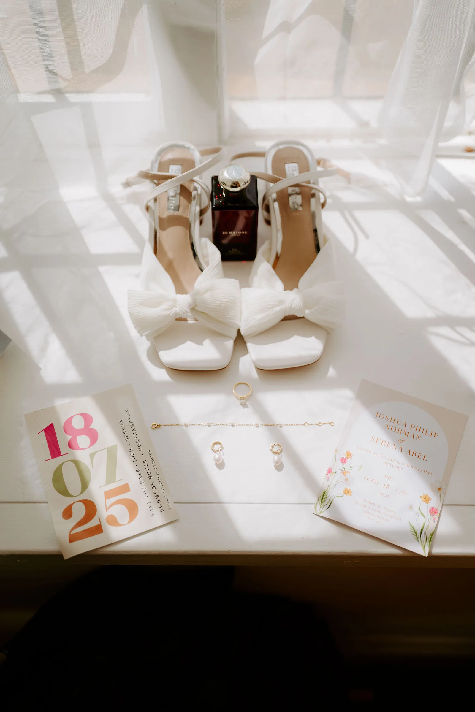 White bridal shoes with bows, a perfume bottle, rings and a printed date card in soft morning window light