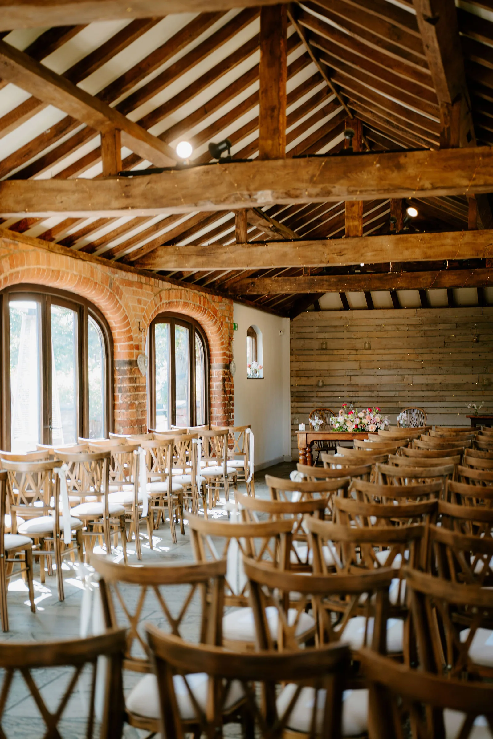 A barn with exposed oak roof beams and brick arched windows, rows of wooden cross-back chairs set out on a flagstone floor facing a small table of flowers, ready for a ceremony