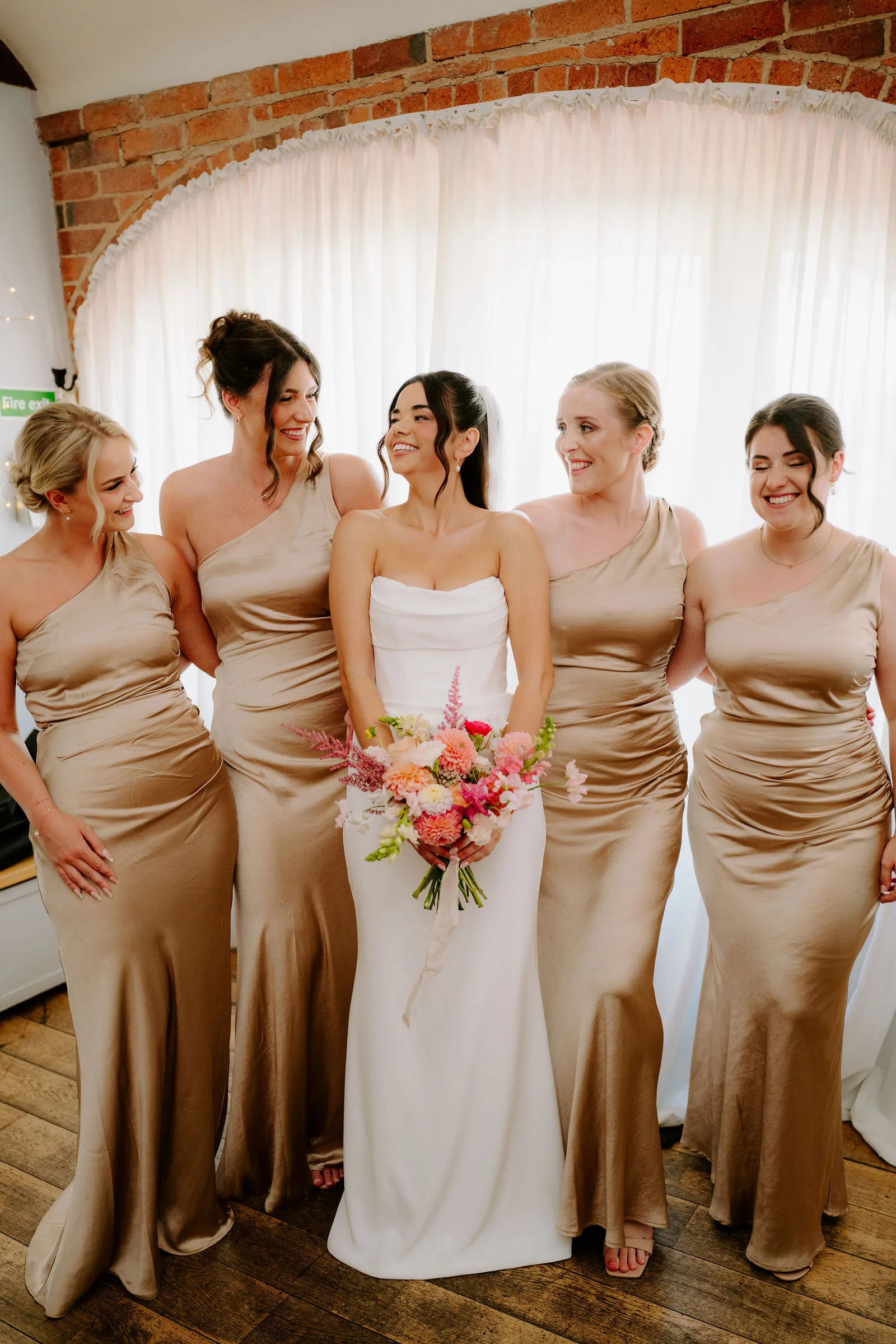A bride in a strapless white dress smiles among four bridesmaids in gold satin dresses, holding a bouquet of pink and orange flowers, in a room with a brick arch hung with white curtains