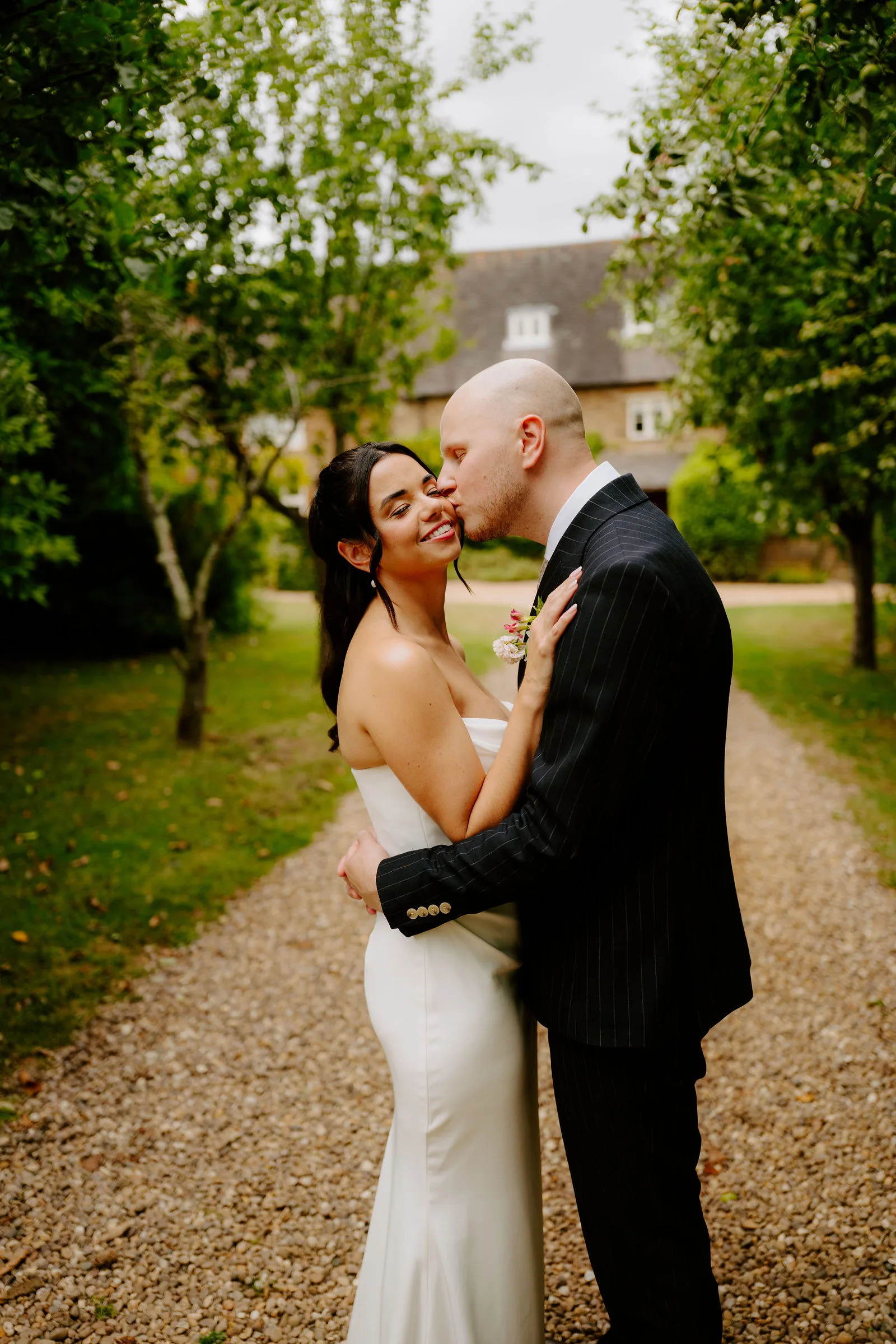 A bride in a strapless white gown smiles as her groom in a dark pinstripe suit kisses her cheek on a gravel path beneath trees, with the stone buildings of Dodmoor House behind them