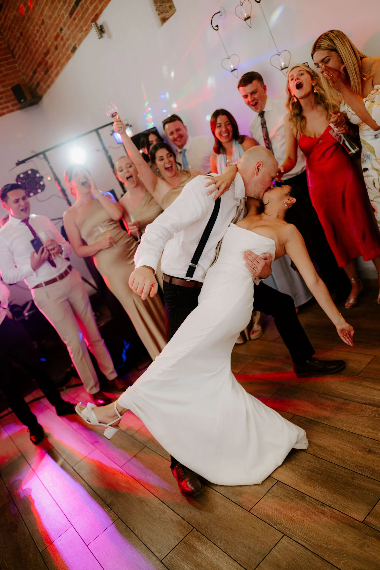 A bride in a white gown is dipped by her groom in a white shirt and braces on a wooden dance floor lit pink, surrounded by cheering guests