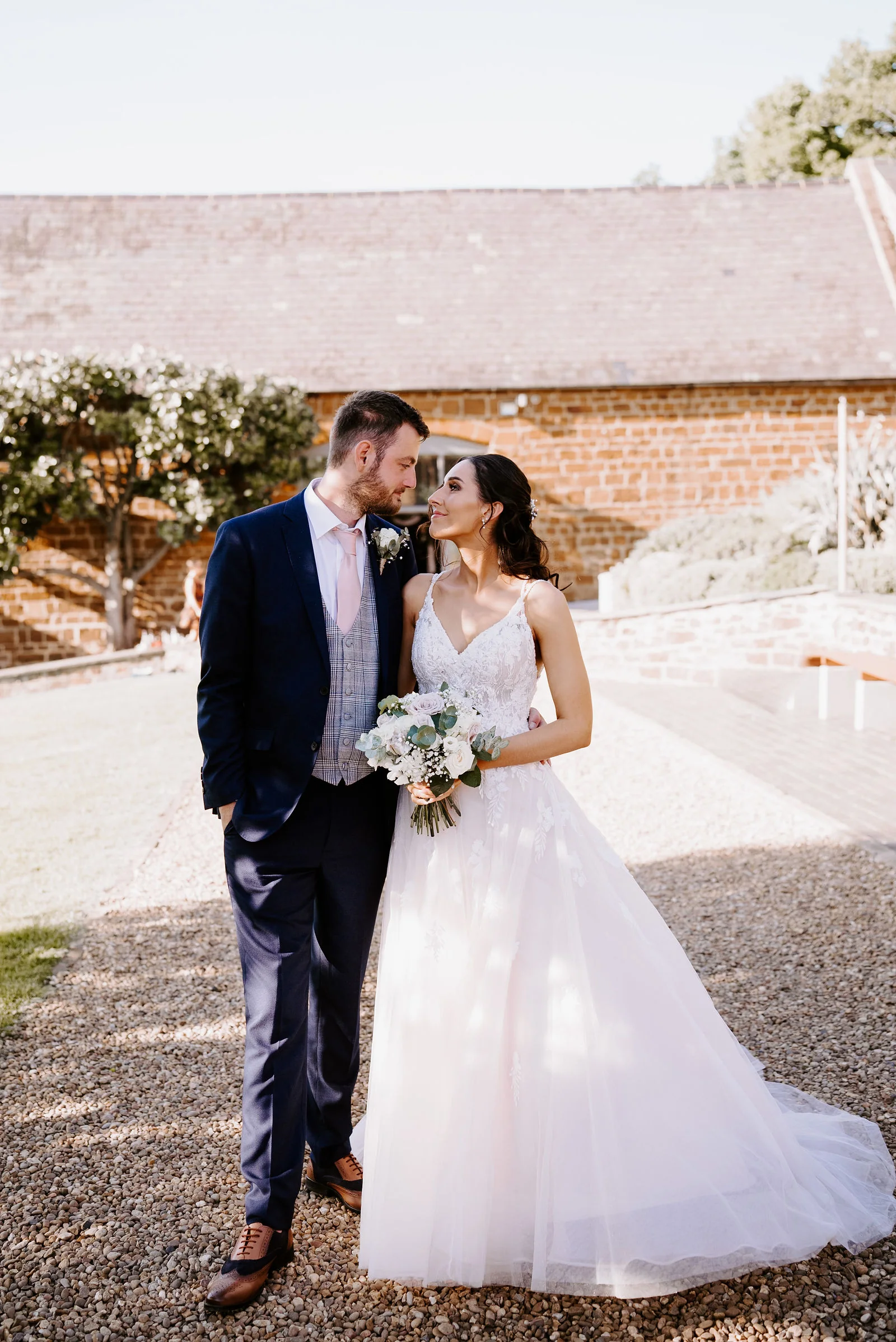 A bride holding a white bouquet and a groom in a navy suit with a grey waistcoat and pink tie stand close on gravel in front of a red ironstone barn with a climbing plant