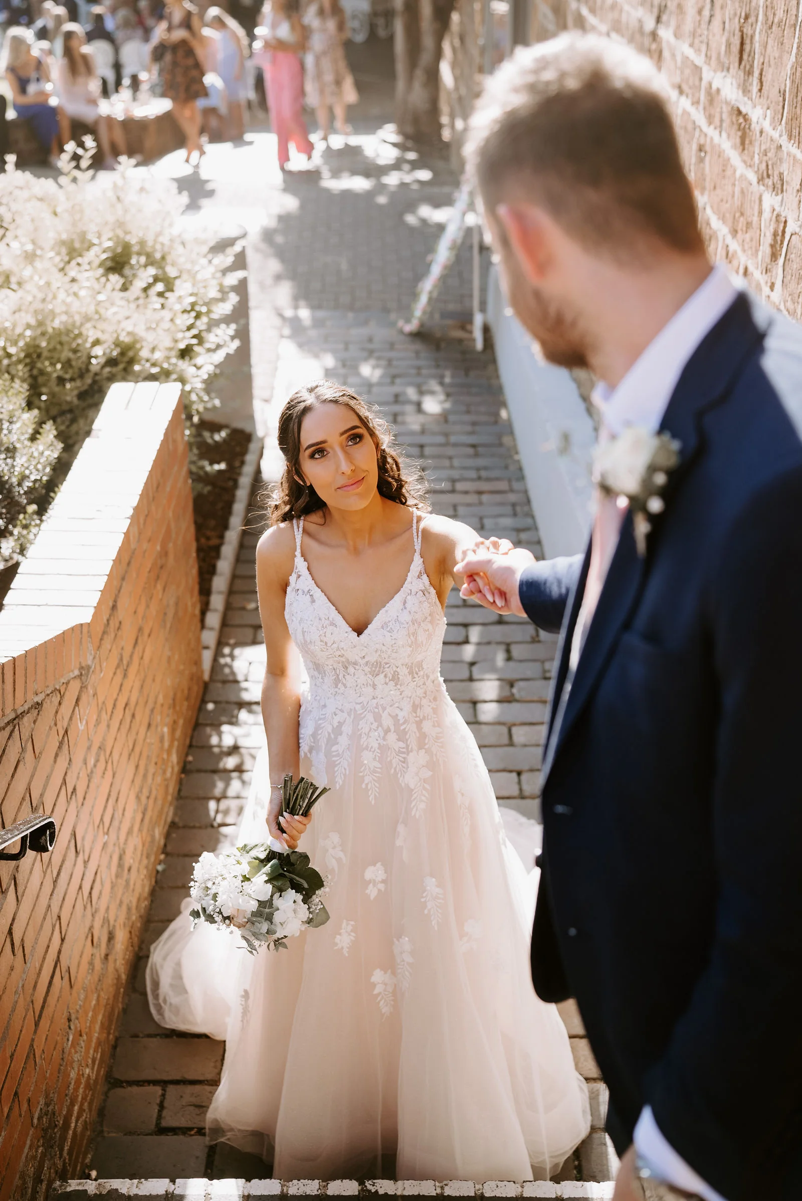 A groom in a navy suit leads his bride in a long tulle gown by the hand along a narrow brick path beside a hedge and a brick wall, with guests gathered at the far end