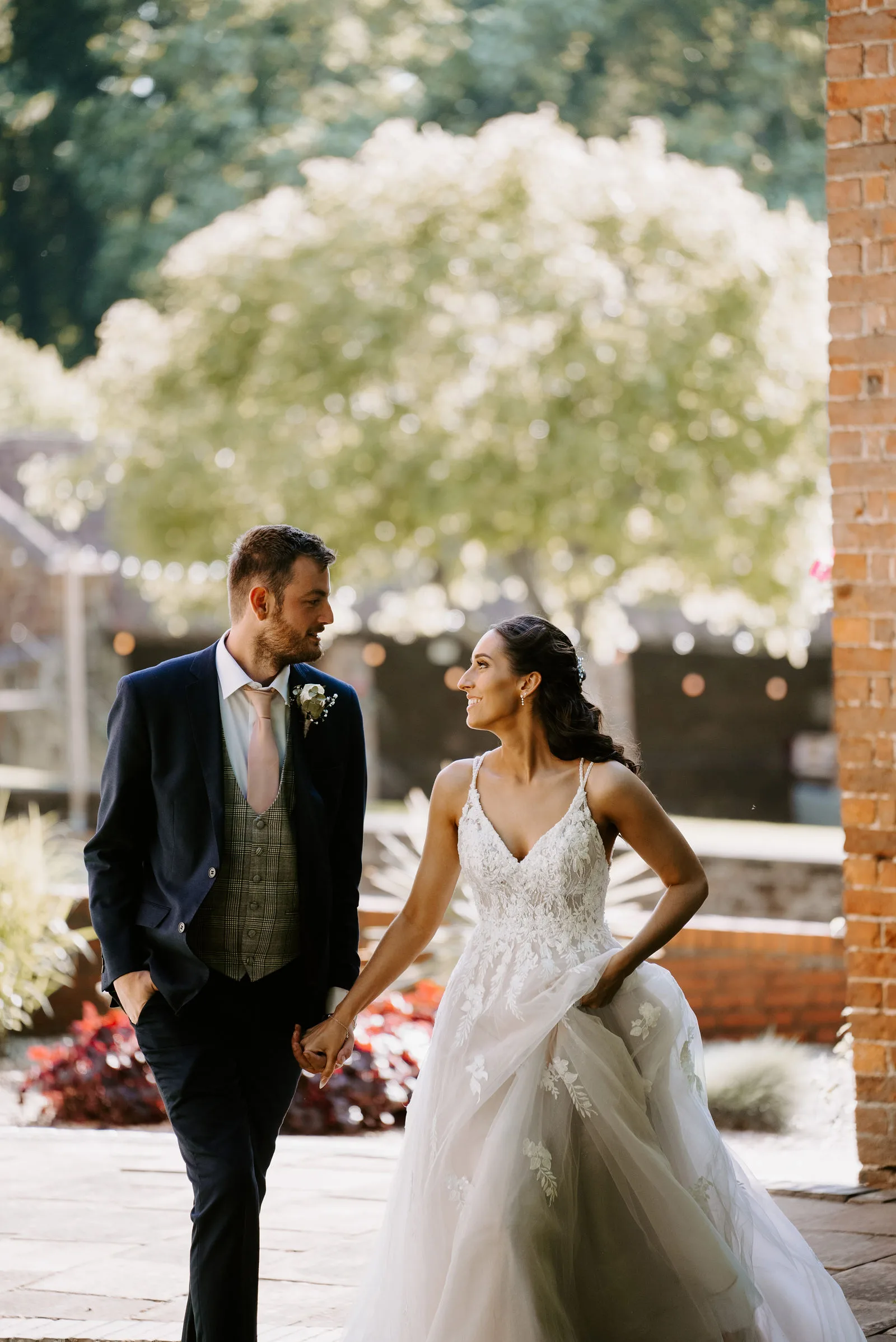A bride in a white gown and a groom in a navy suit walk hand in hand and smile at each other along a brick-paved path beside a brick wall, with a tree in blossom and strings of lights behind them
