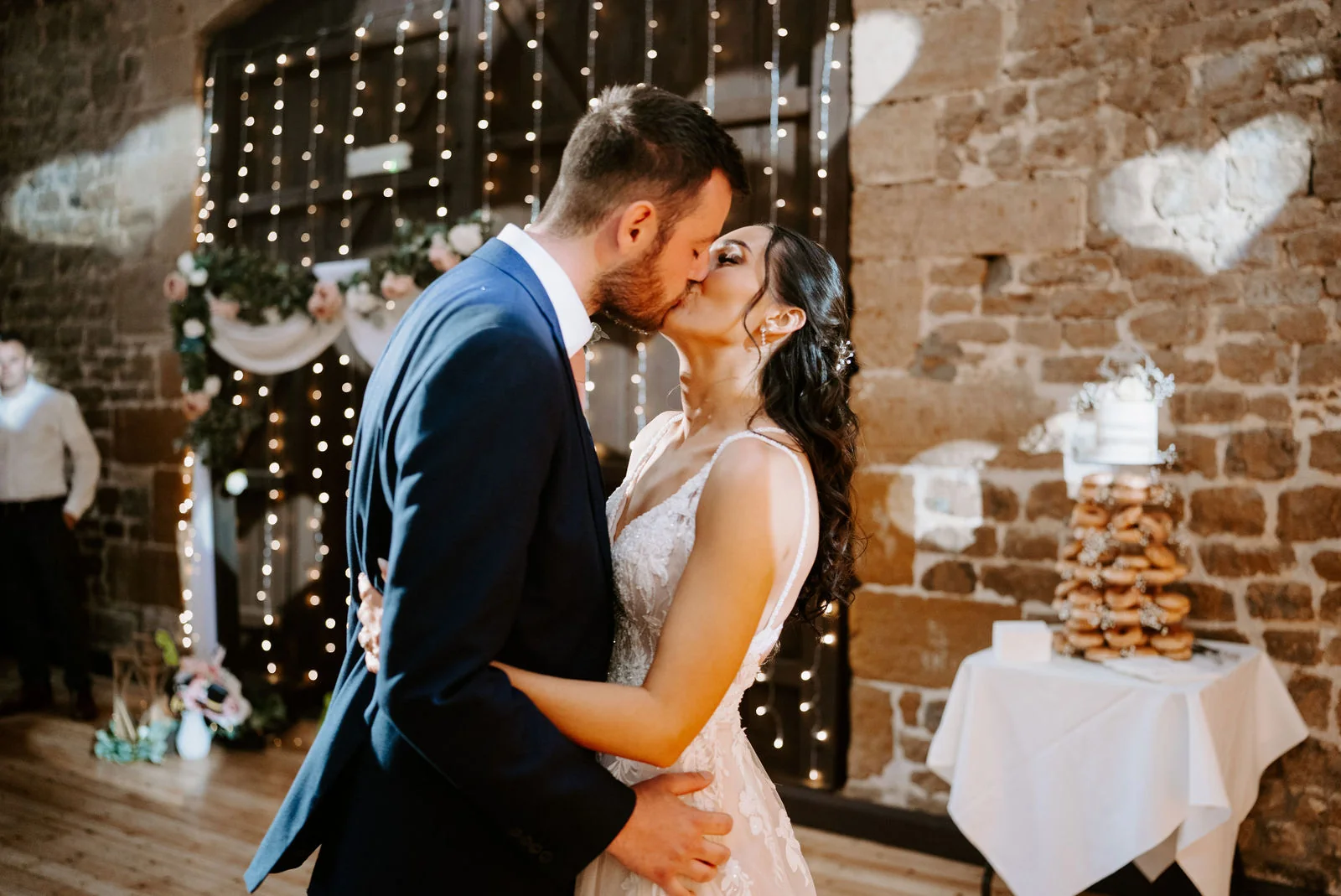 A bride and groom kiss in a barn in front of a curtain of fairy lights, with a tall stack of doughnuts on a table beside them