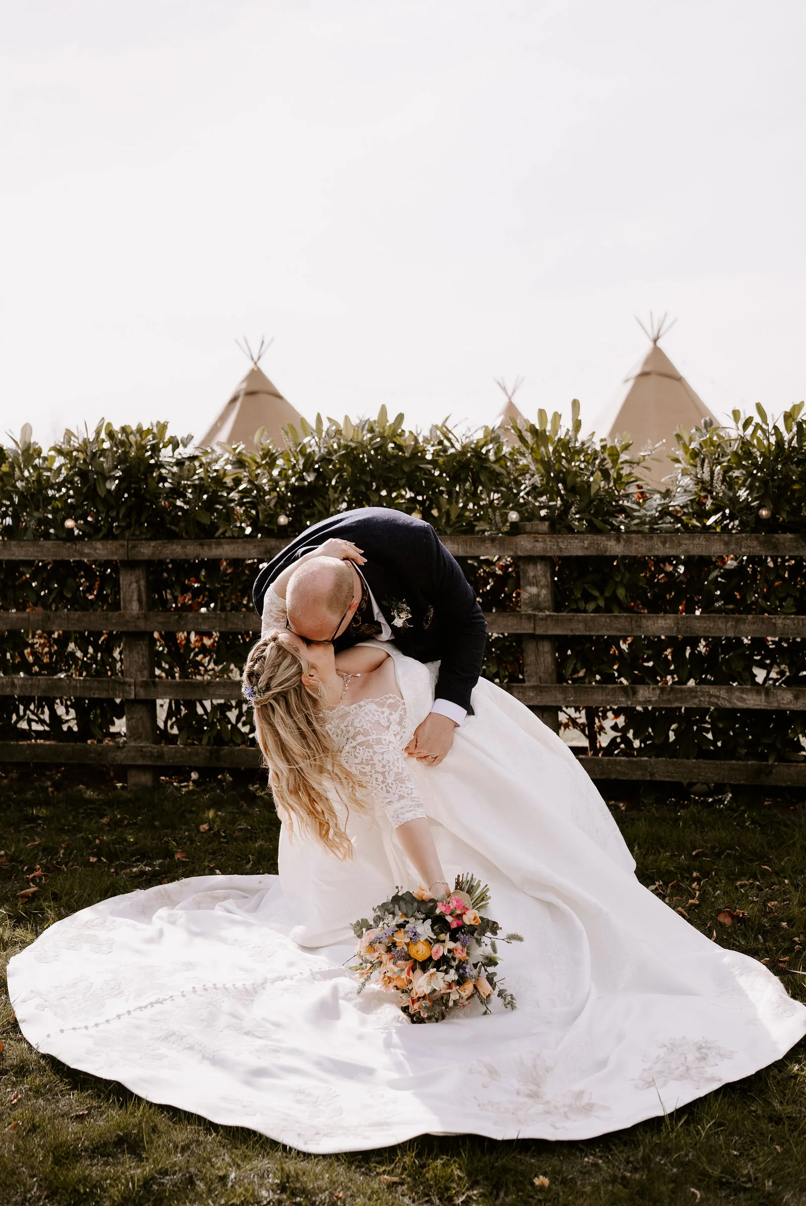 A groom in a dark suit dips a bride in a full satin gown holding a pink and orange bouquet to kiss her on grass in front of a wooden fence and hedge, with the tips of tipi tents beyond