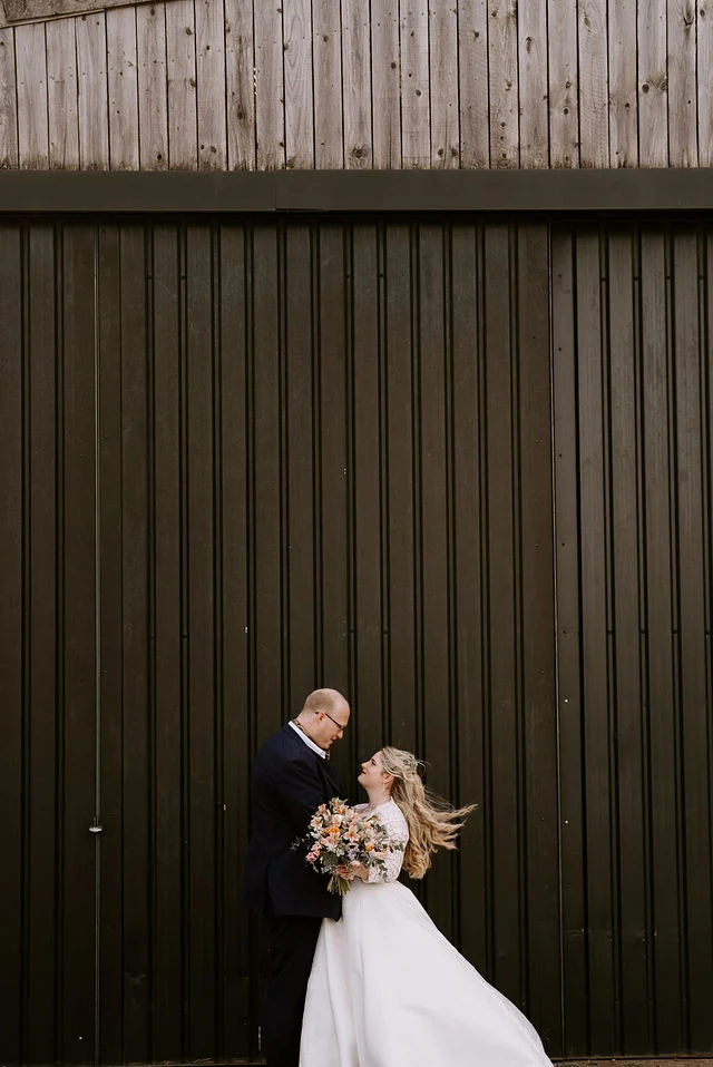Becky and Rob standing together against a tall timber barn wall