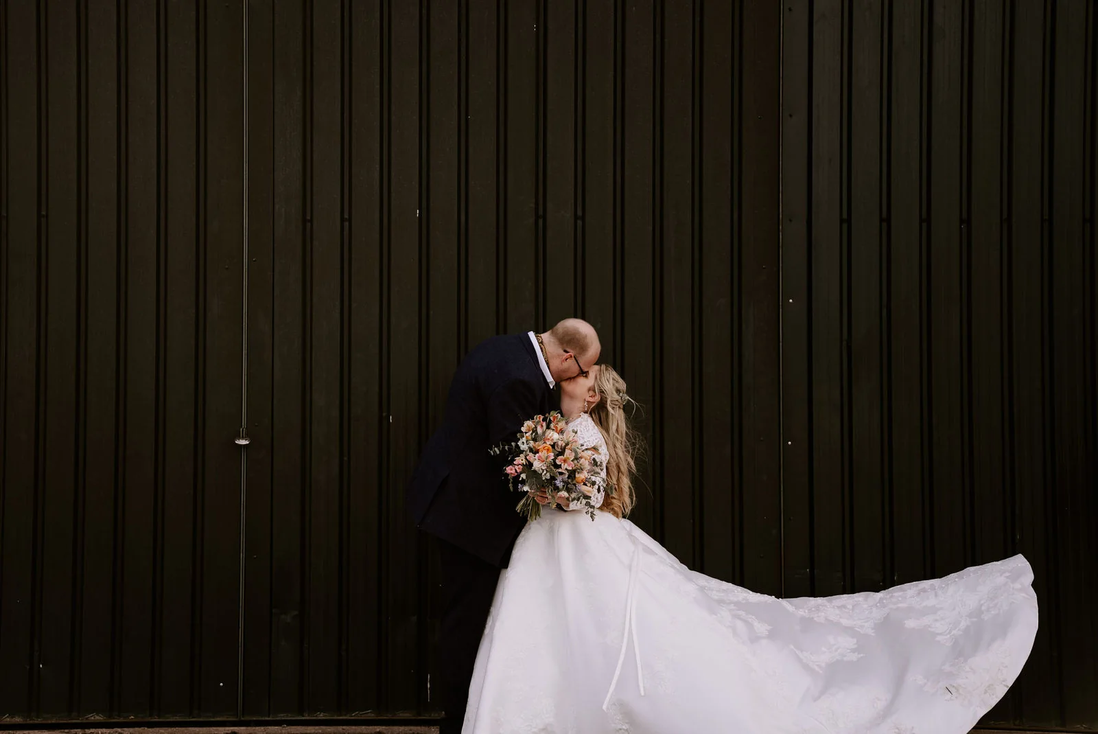 A couple kiss in front of a tall dark timber barn wall, the bride’s long train spread out behind her