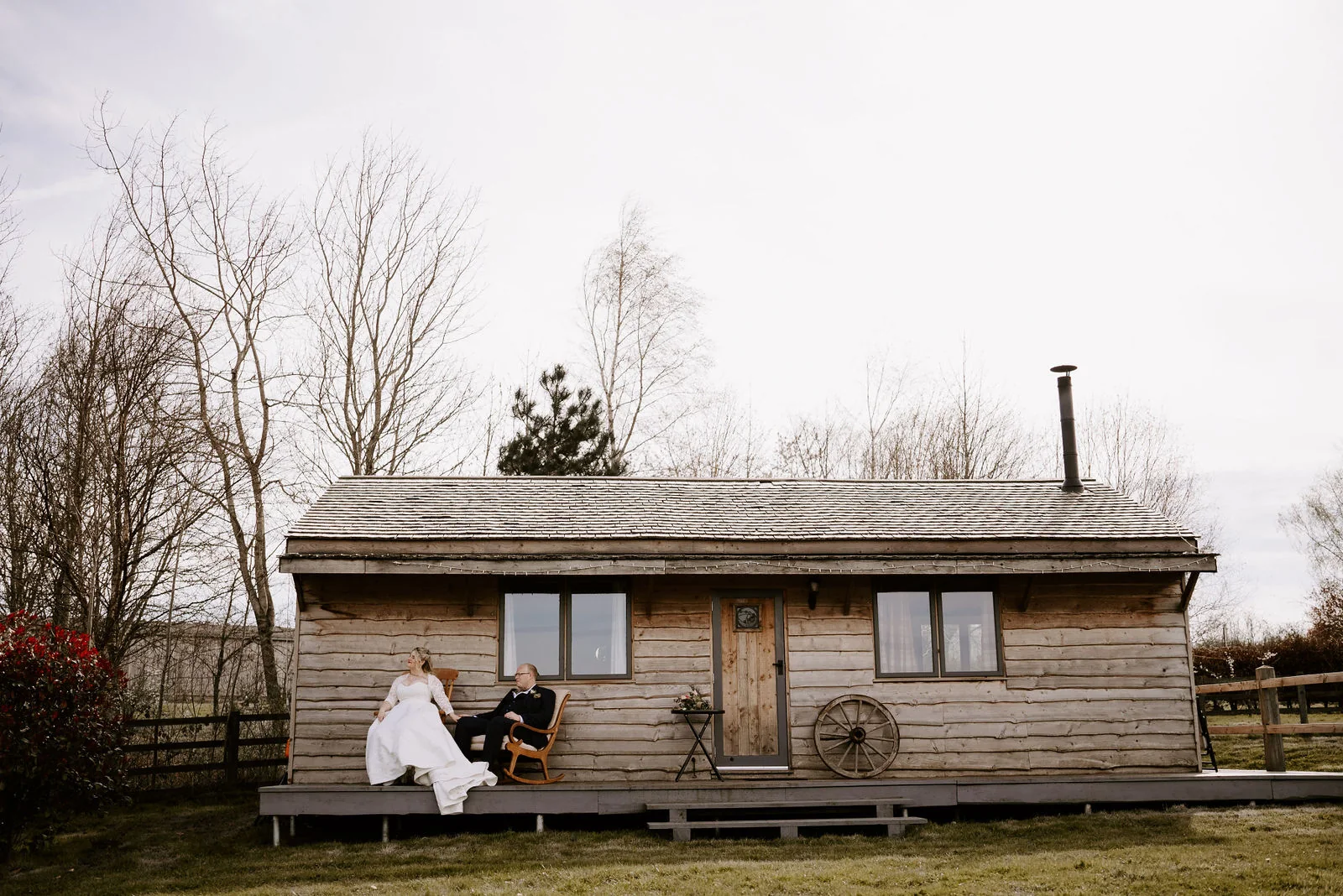 A bride and a groom sit on rocking chairs on the decking of a small weathered timber cabin with a wagon wheel by the door, with bare trees behind