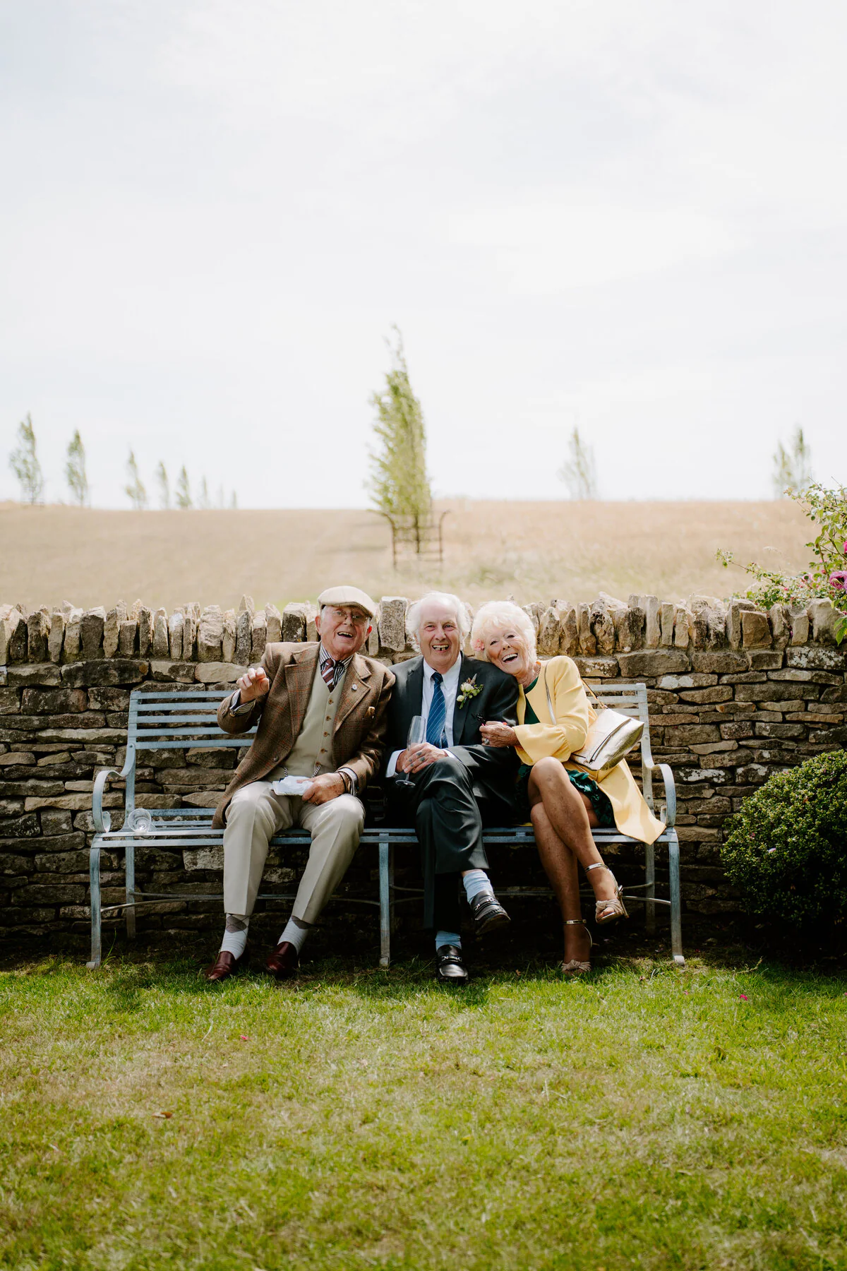 Three wedding guests laughing together on a bench beside a stone wall.