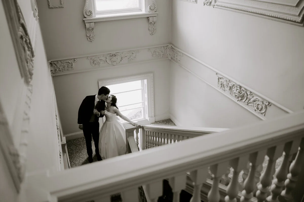 Black and white photograph of newlyweds sharing a kiss on an elegant staircase.