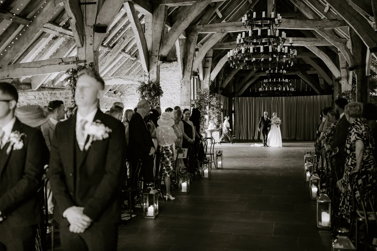 Black and white photograph of a bride and her father walking down a lantern-lit aisle beneath the timber roof and chandelier of a barn, with guests on either side.